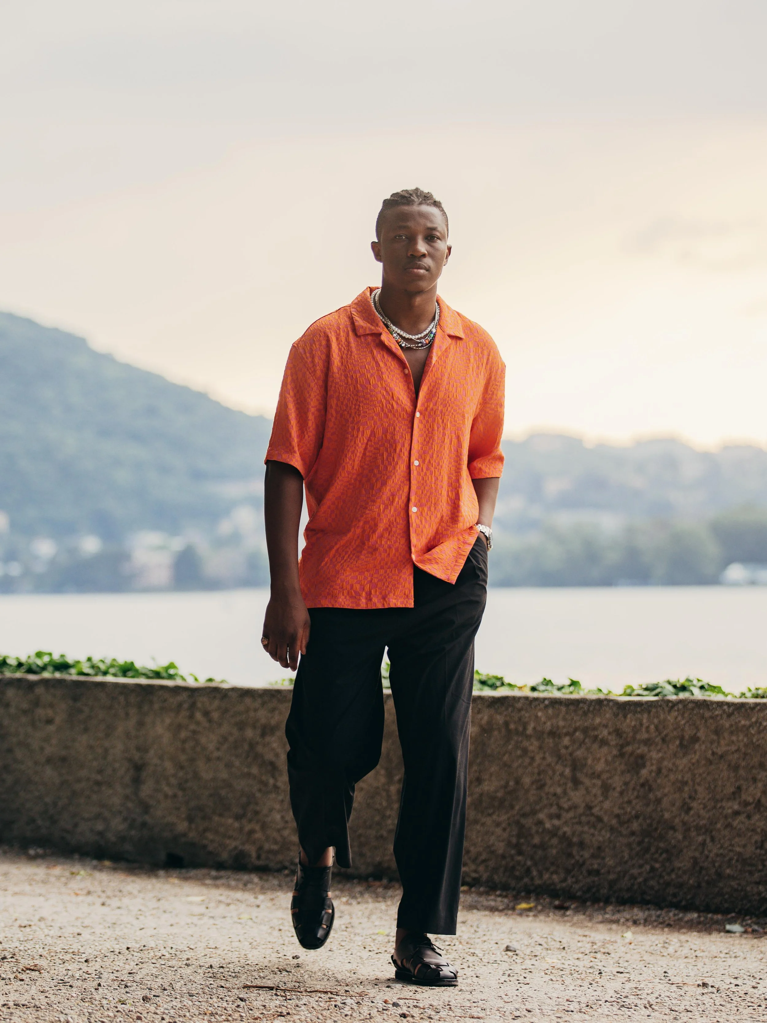 A young man walking outdoors near a body of water, wearing an orange patterned short-sleeve shirt, black pants, black shoes, and layered necklaces with a watch on his left wrist.