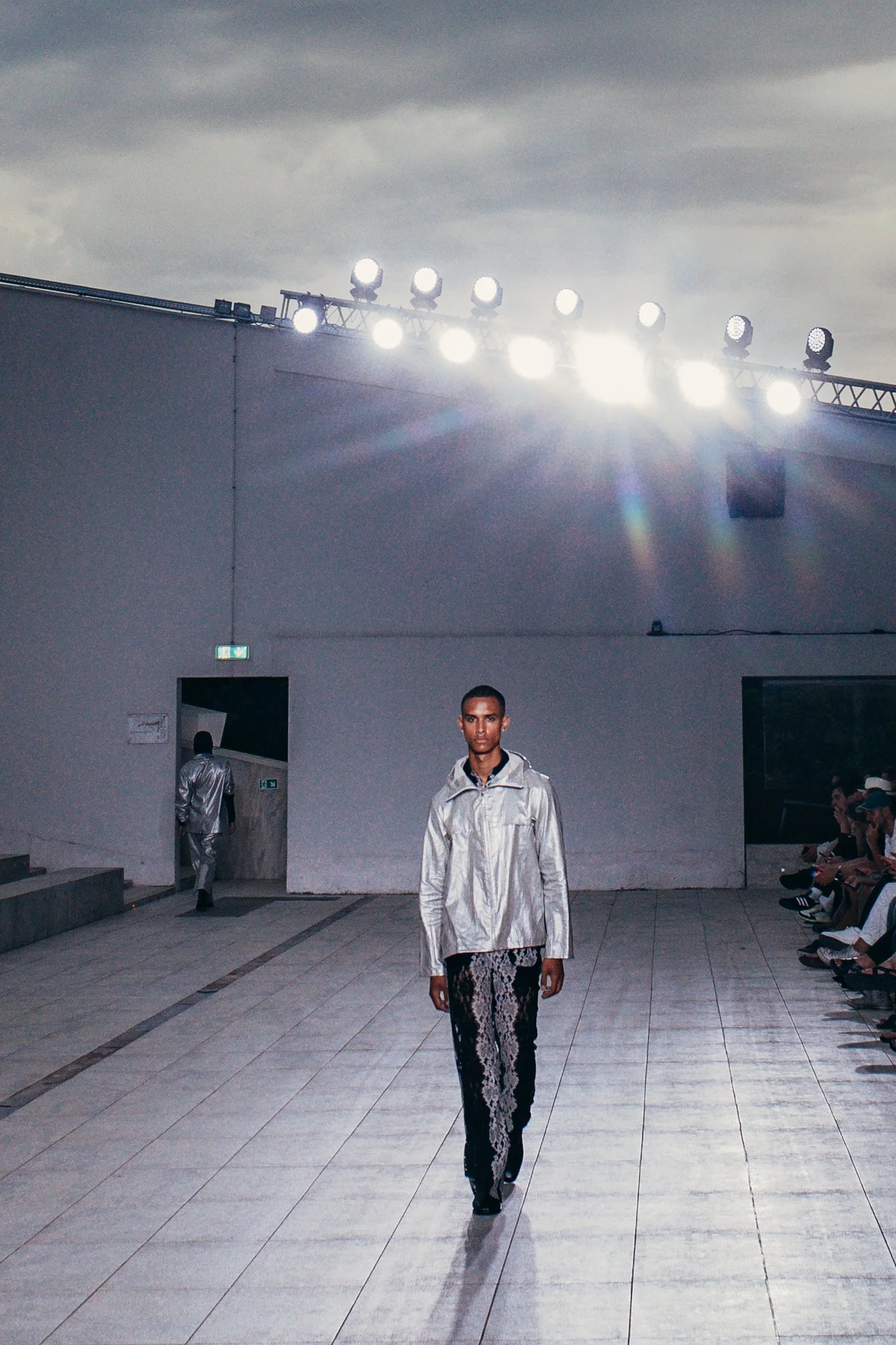 Male model walking on a runway during a fashion show, with a white wall, audience, and stage lights in the background.