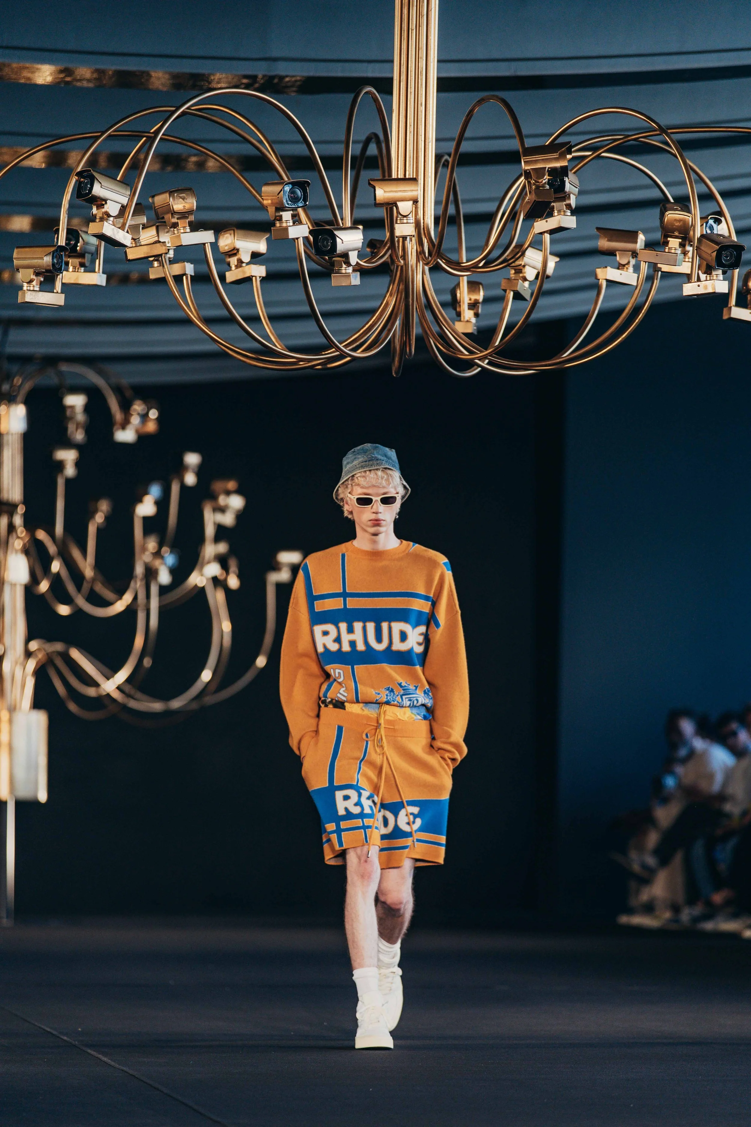 Male model walking on runway in a mustard yellow and blue outfit with text 'RHODE' and oversized sunglasses, under a large gold chandelier in a fashion show.