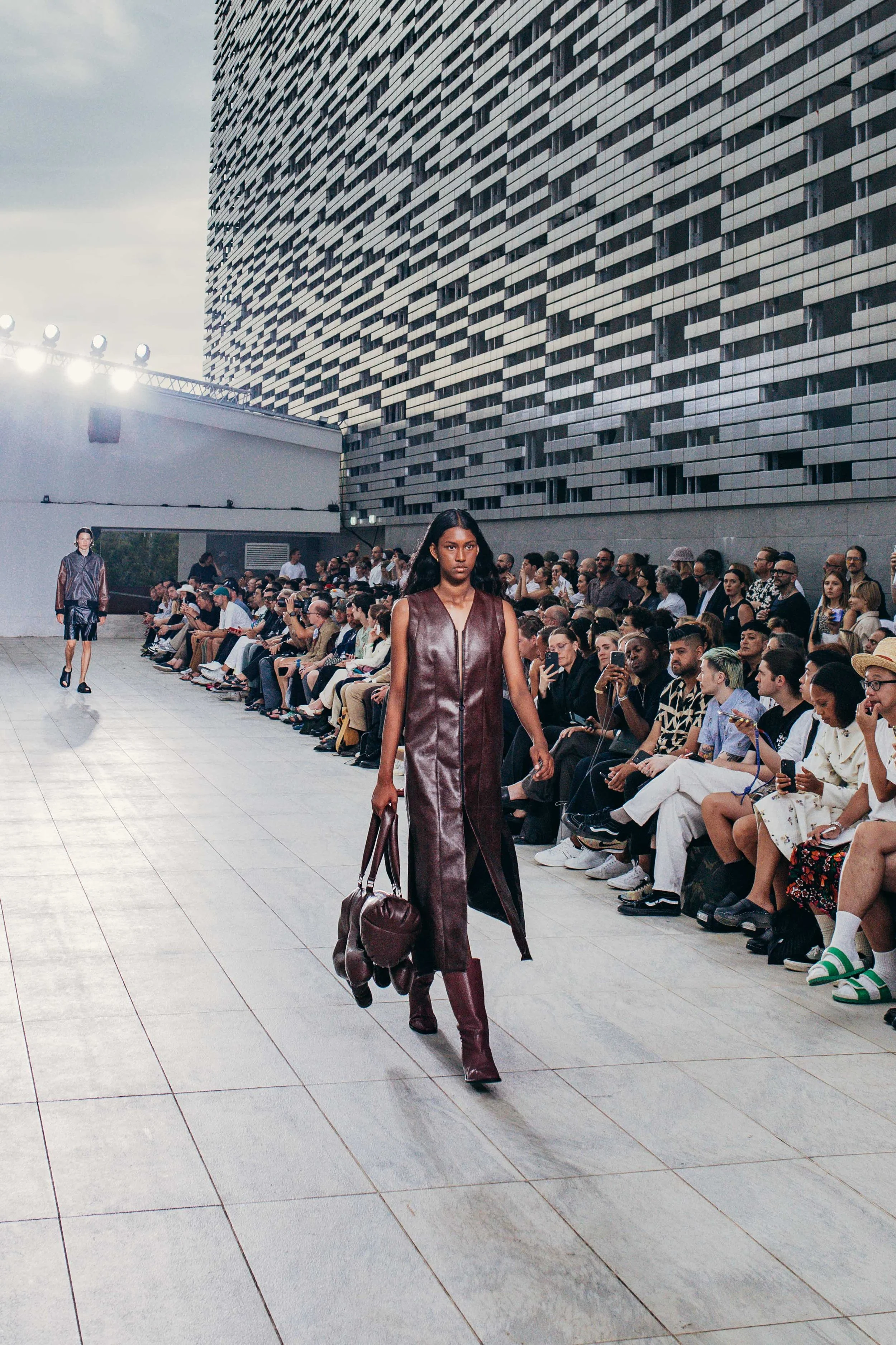 Model walking on runway at a fashion show with audience seated on both sides, urban setting with modern building backdrop.