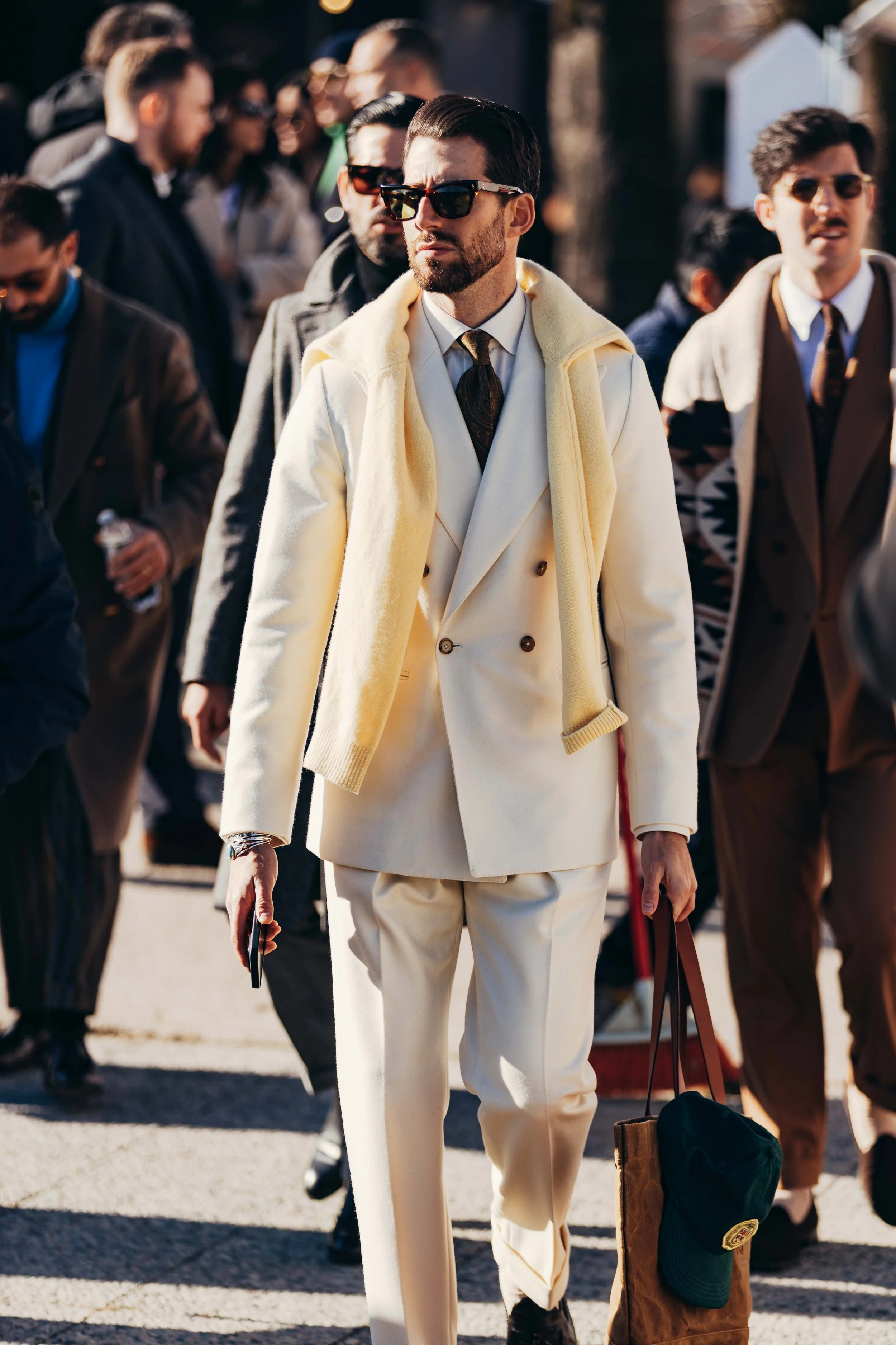 A stylish man walking outdoors in a crowd, wearing sunglasses, a white suit with a double-breasted jacket, and holding a phone and a bag, with others dressed in formal and casual attire around him.