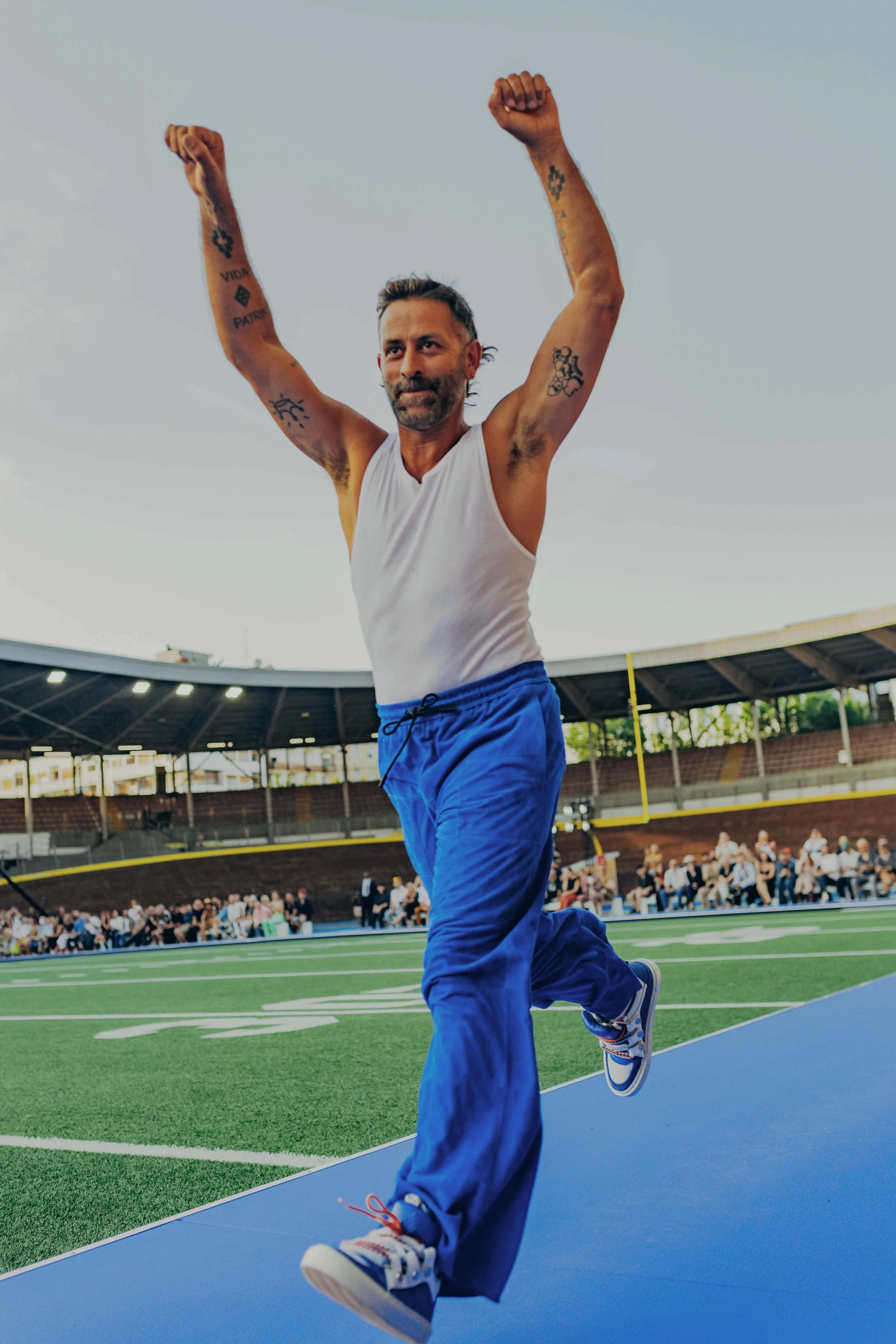 A man with tattoos running on a football field, arms raised in celebration, with an audience seated in the background.