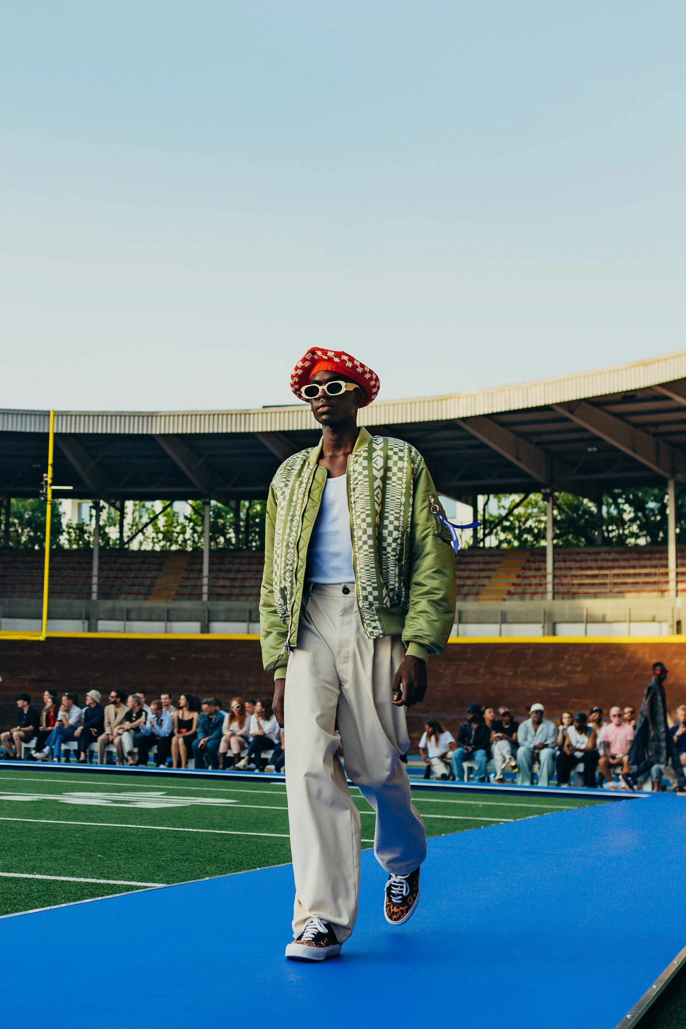 A person walking on a runway at a stadium watching fashion show, wearing green jacket, white pants, leopard print sneakers, red hat, and white sunglasses.