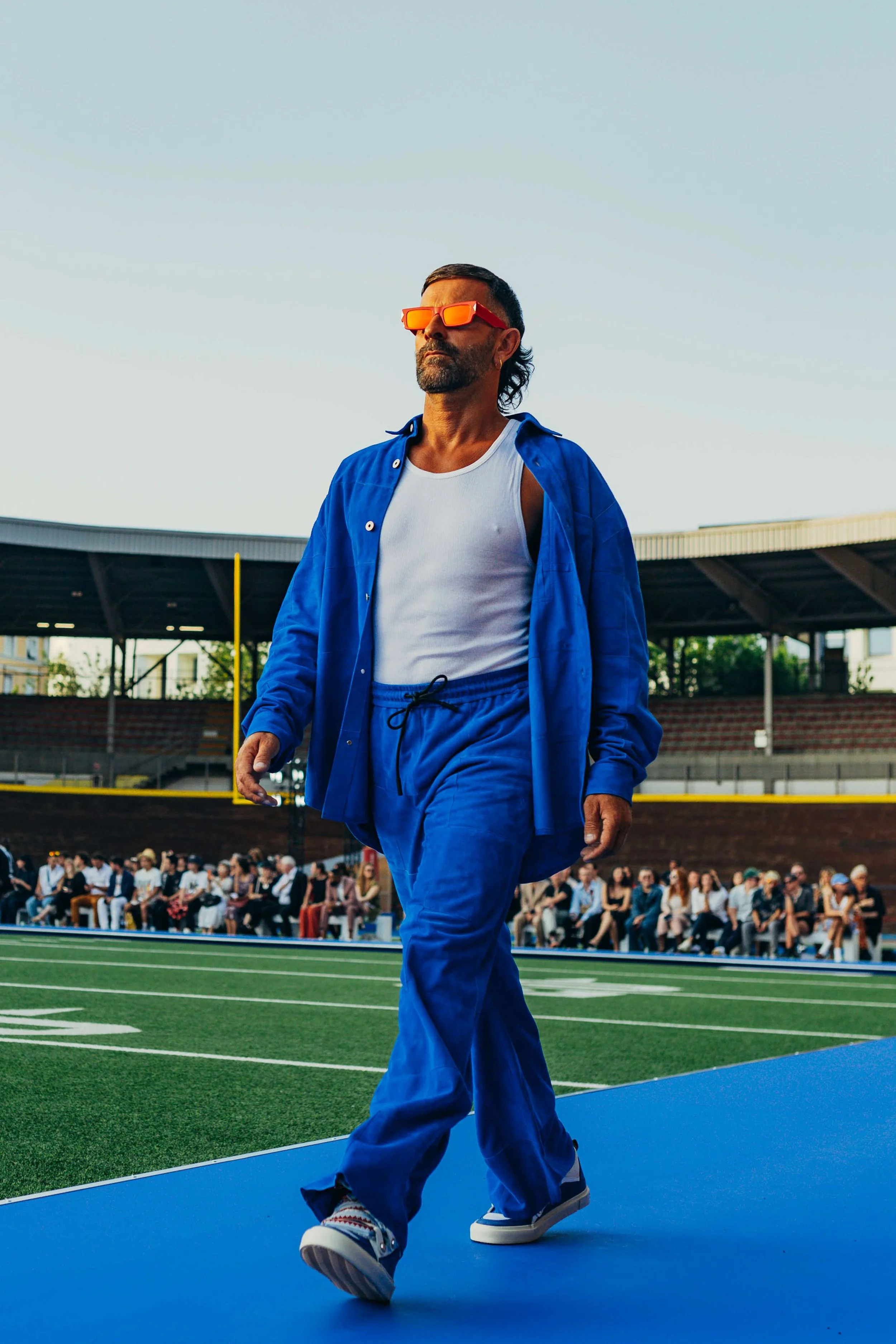 A man with sunglasses and a beard walking on a runway at an outdoor event with a sports stadium in the background, wearing a blue jacket and pants, white tank top, and sneakers.