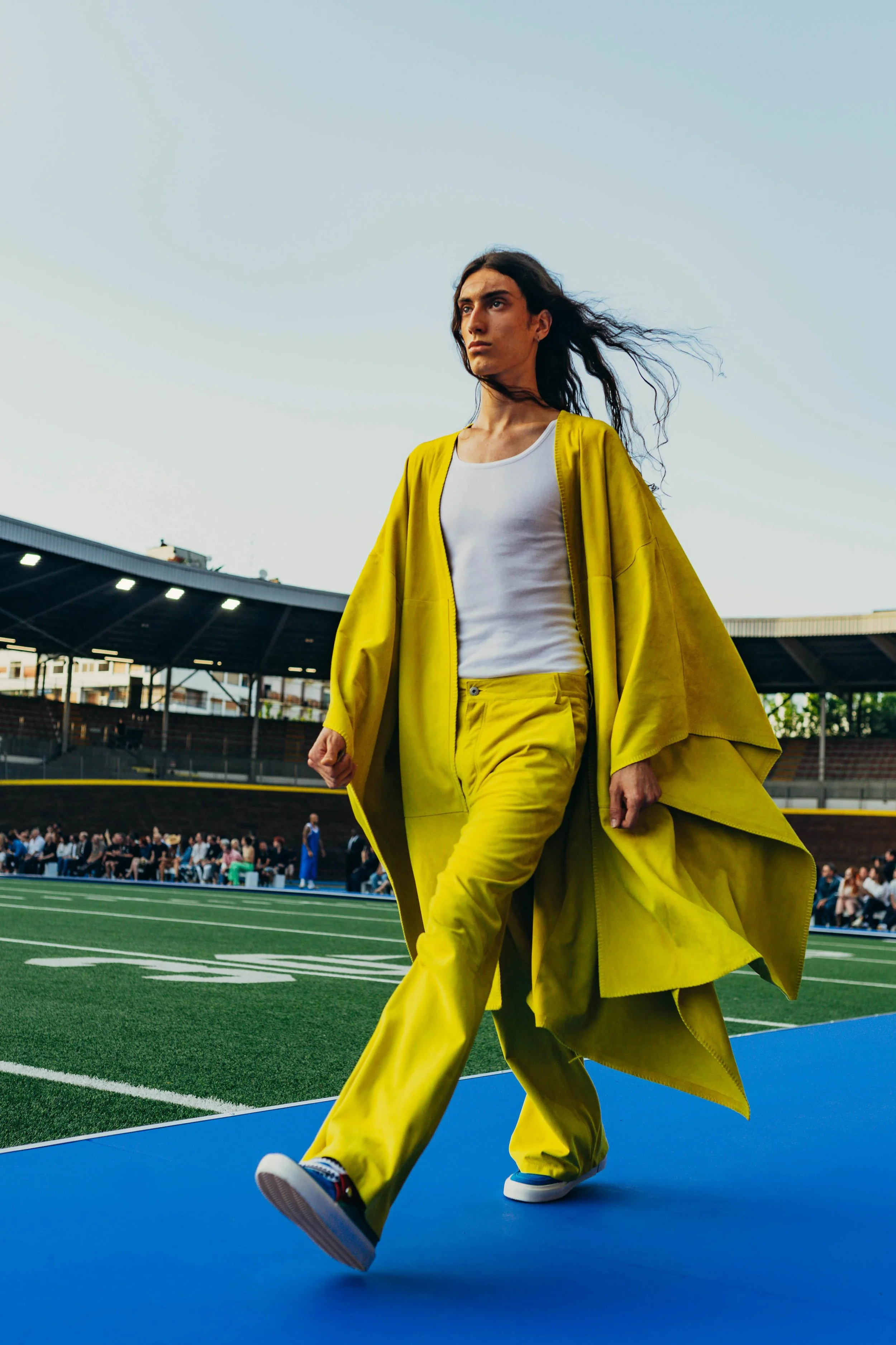 A woman in a yellow coat and matching yellow pants walking on a blue track in a stadium with an audience in the background.