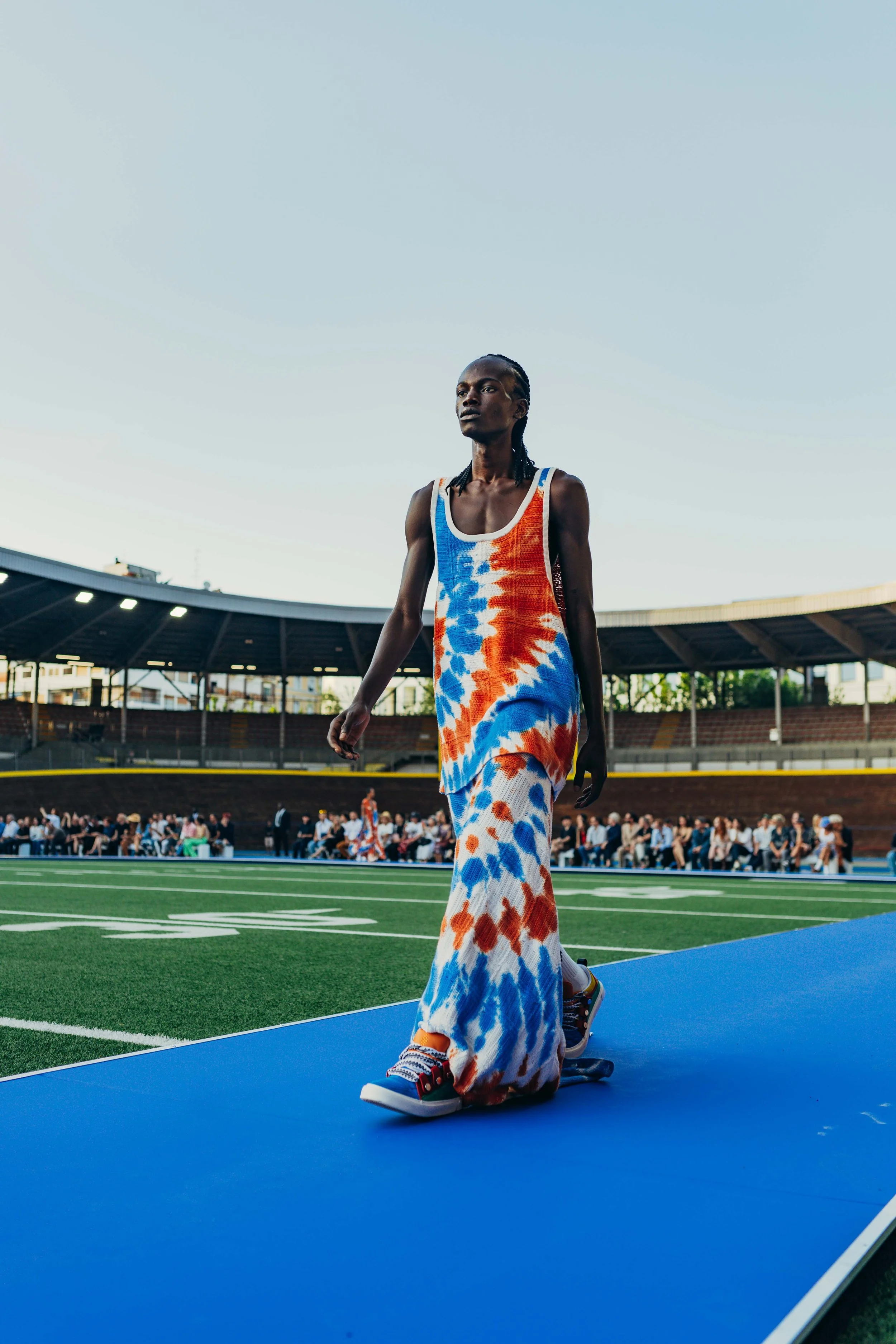 A woman walking on a blue runway at an outdoor stadium, wearing a tie-dye dress and sneakers, with a crowd seated in the background.