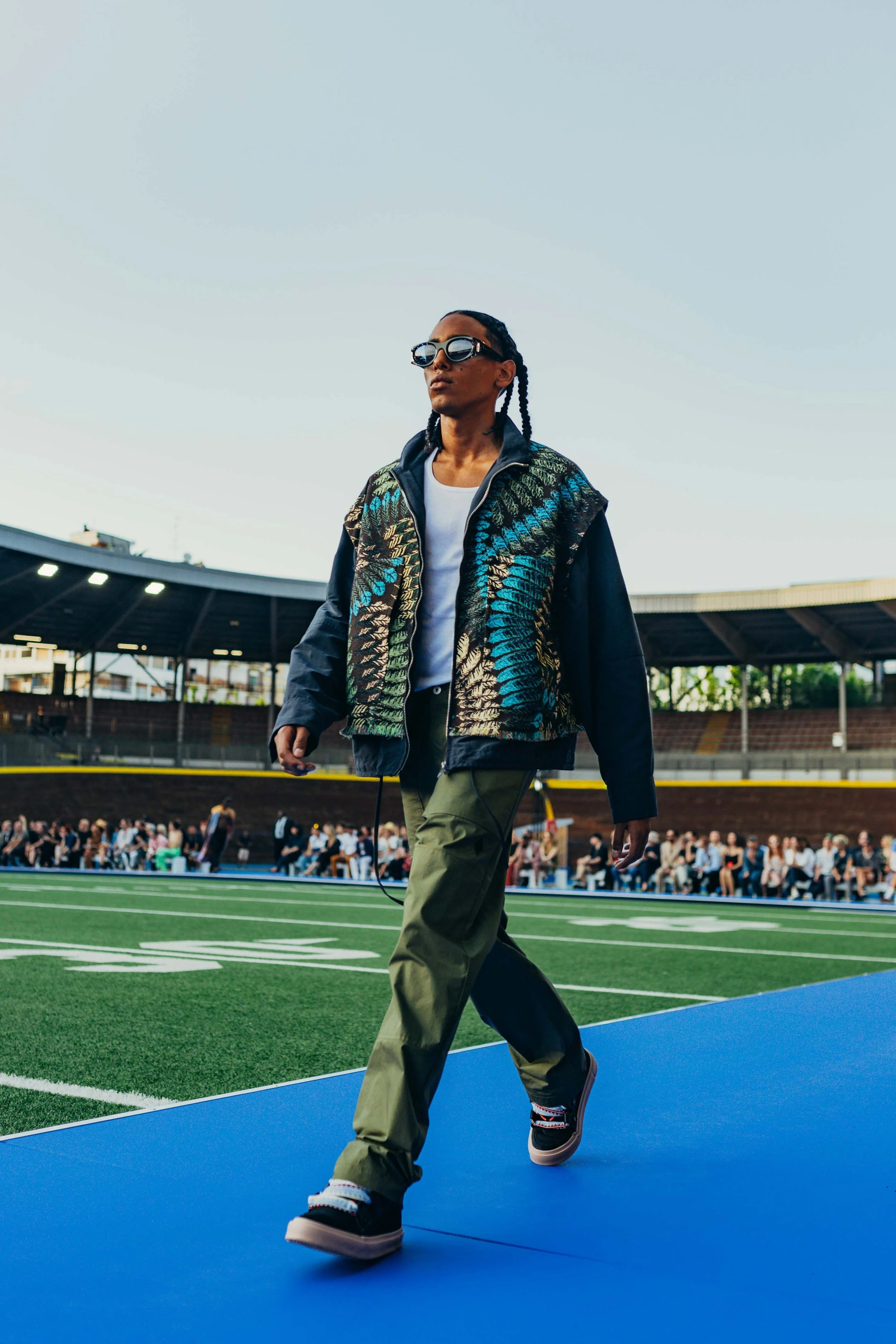 A person walking on a runway outside a stadium, wearing sunglasses, a patterned jacket, white shirt, and cargo pants, with a crowd seated in the background.