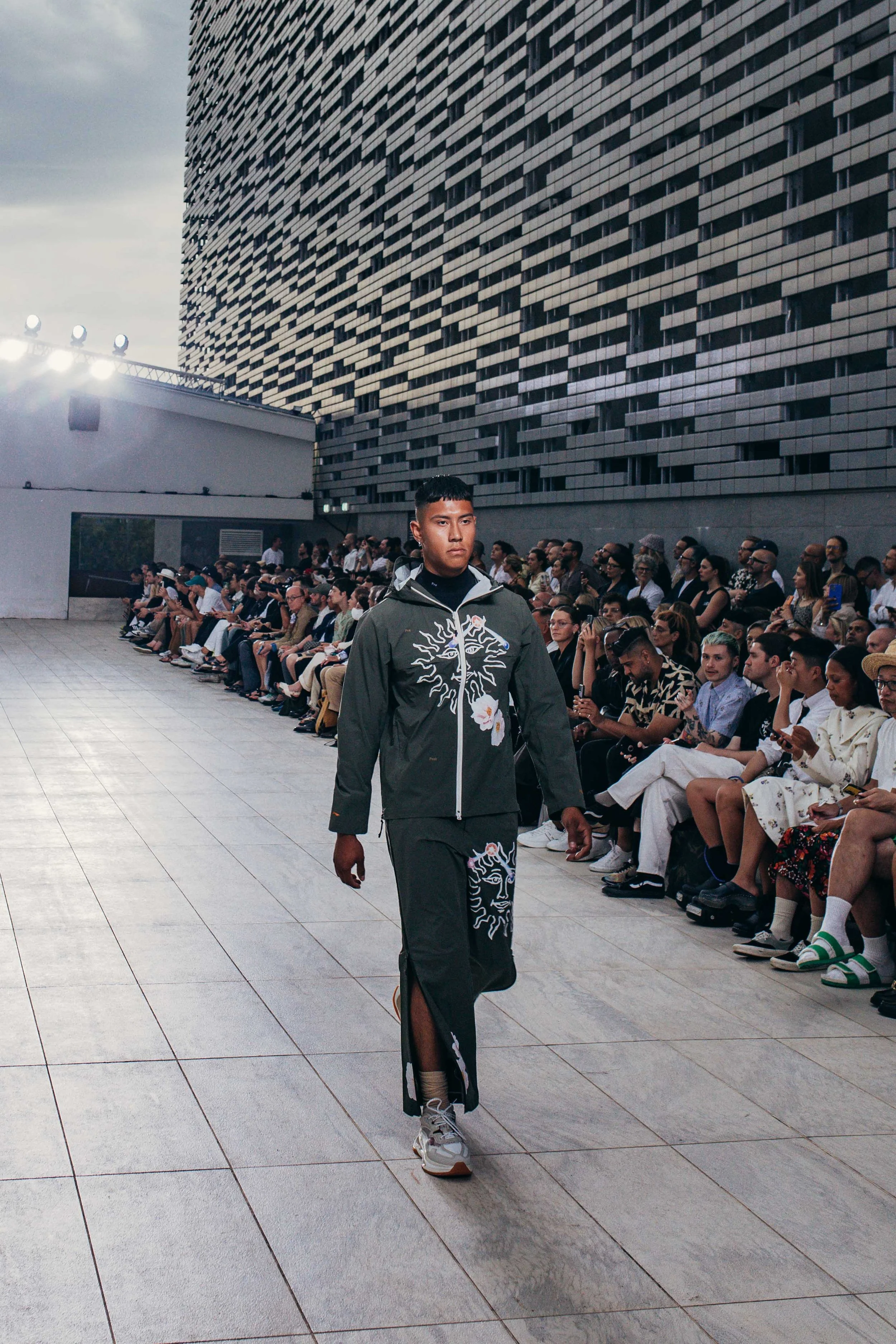 A male model walks on an outdoor runway during a fashion show, with a large audience seated on either side and a modern building in the background.