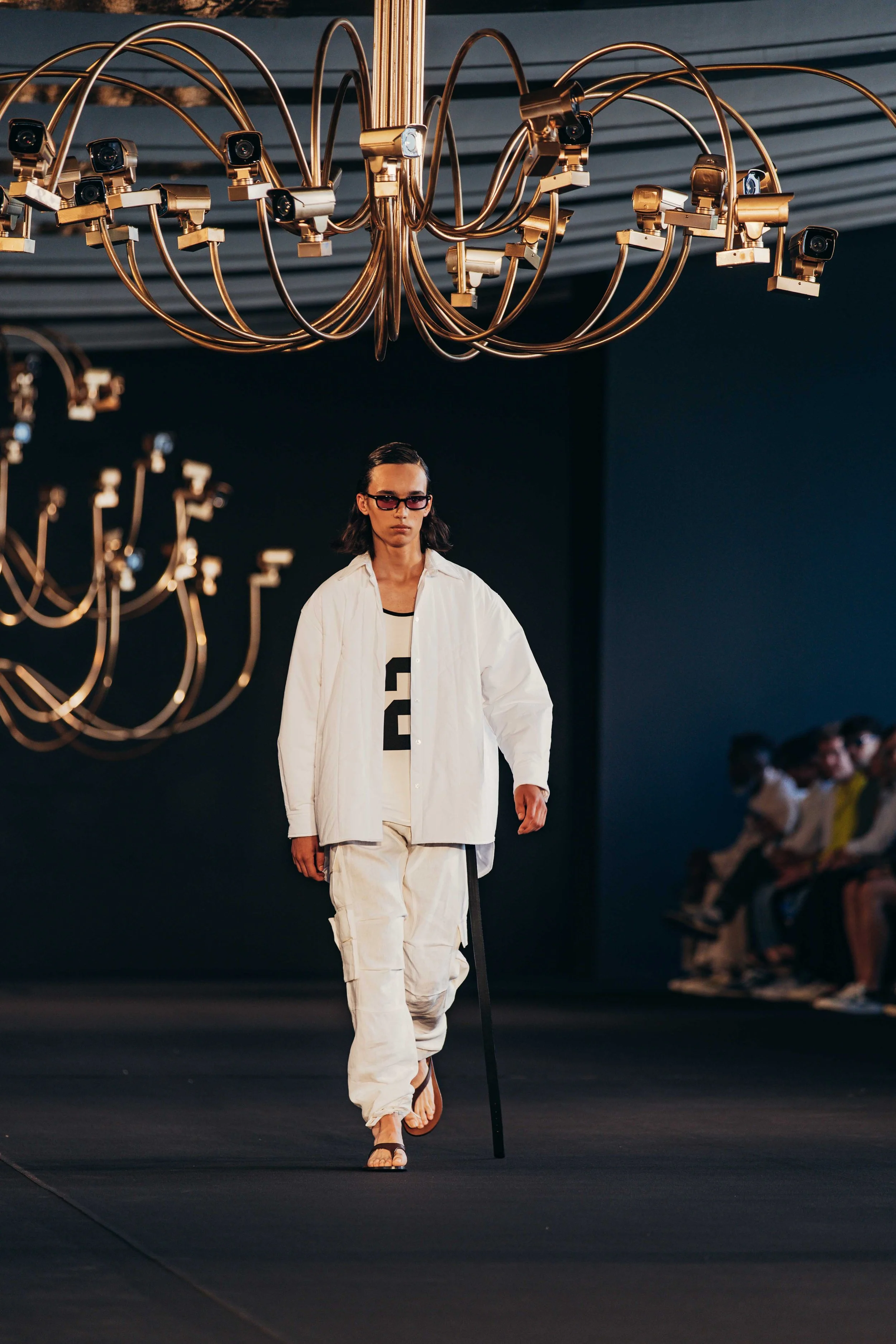 Fashion model walking down runway wearing white layered outfit with black accents, dark sunglasses, and holding a stick, in a dark room with large gold chandeliers overhead.