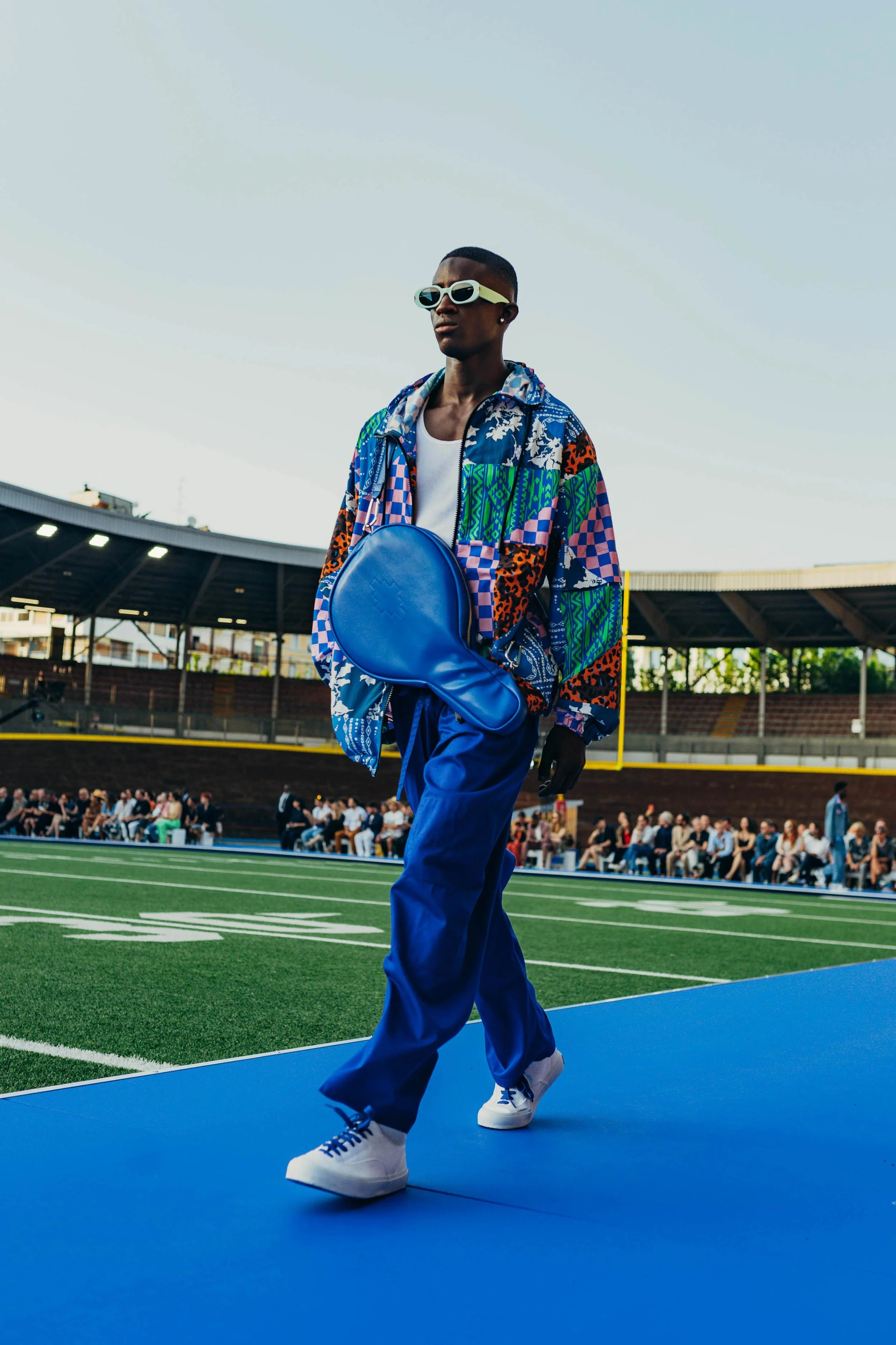 A stylish young man in colorful patterned clothing walking on a blue path at a sports stadium, holding a blue tennis bag, with spectators in the background.