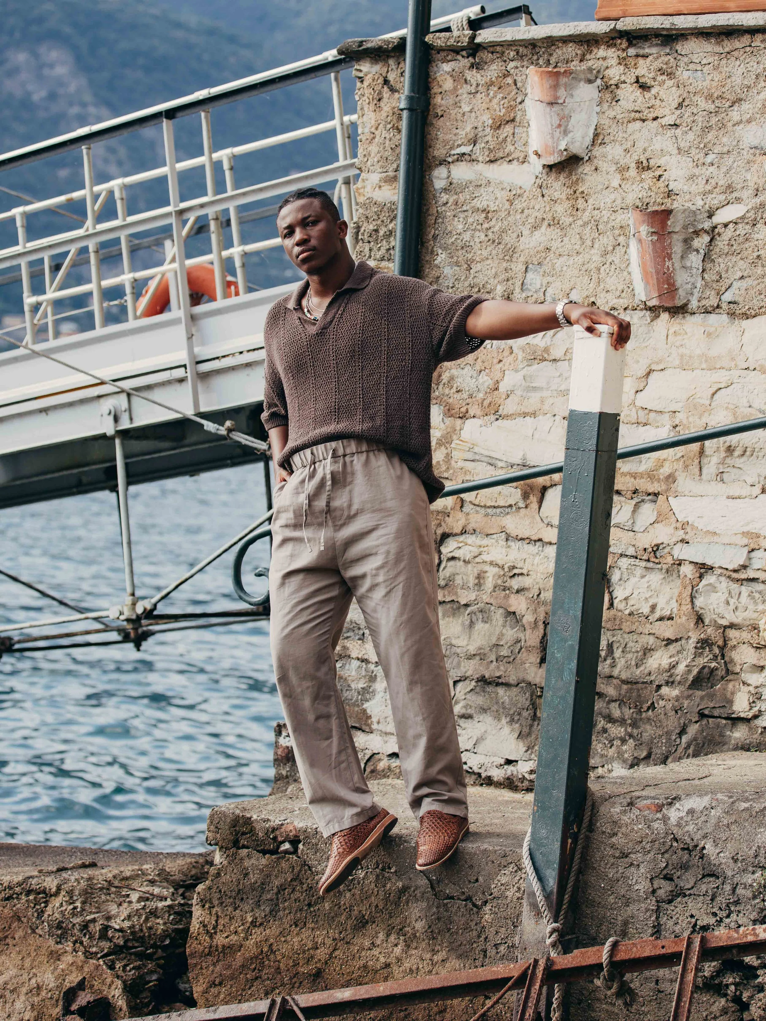 A person standing on rocks by the water, wearing a brown sweater, beige pants, and brown shoes, with a stone wall and boat dock in the background.