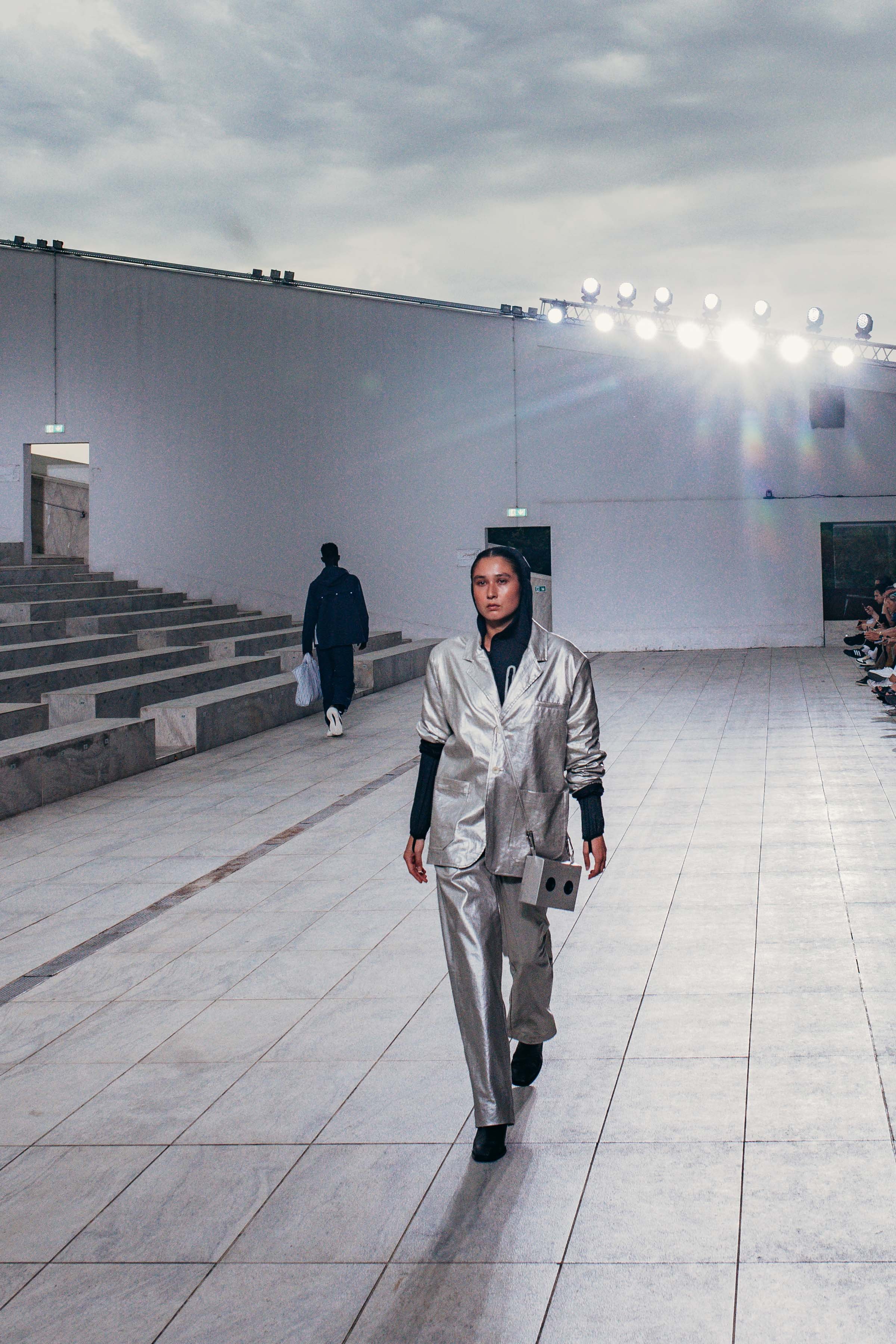 A woman walking on a runway during a fashion show, wearing metallic silver suit and carrying a small bag, with a few people seated on the right and a man walking away in the background.