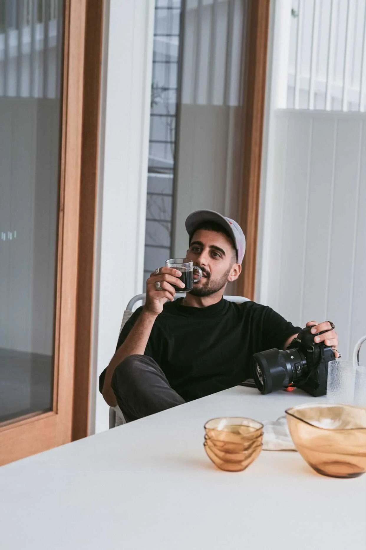 A man sitting at a dining table, drinking from a glass of dark-colored beverage, holding a camera in his left hand, wearing a black t-shirt and a gray cap, with light-colored walls, windows, and bowls on the table.