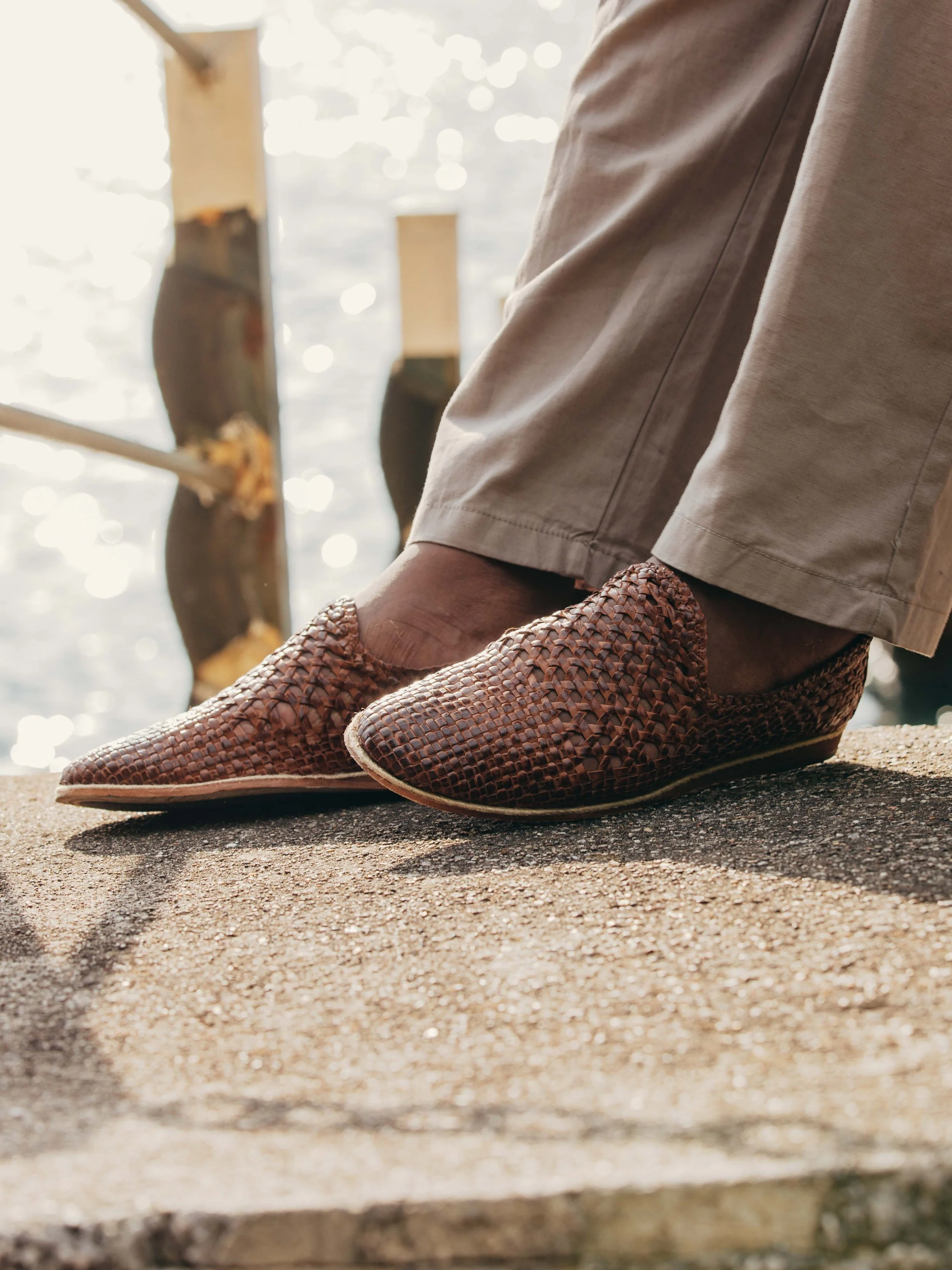 Person wearing woven leather shoes standing on a concrete surface near water, with a wooden post in the background.