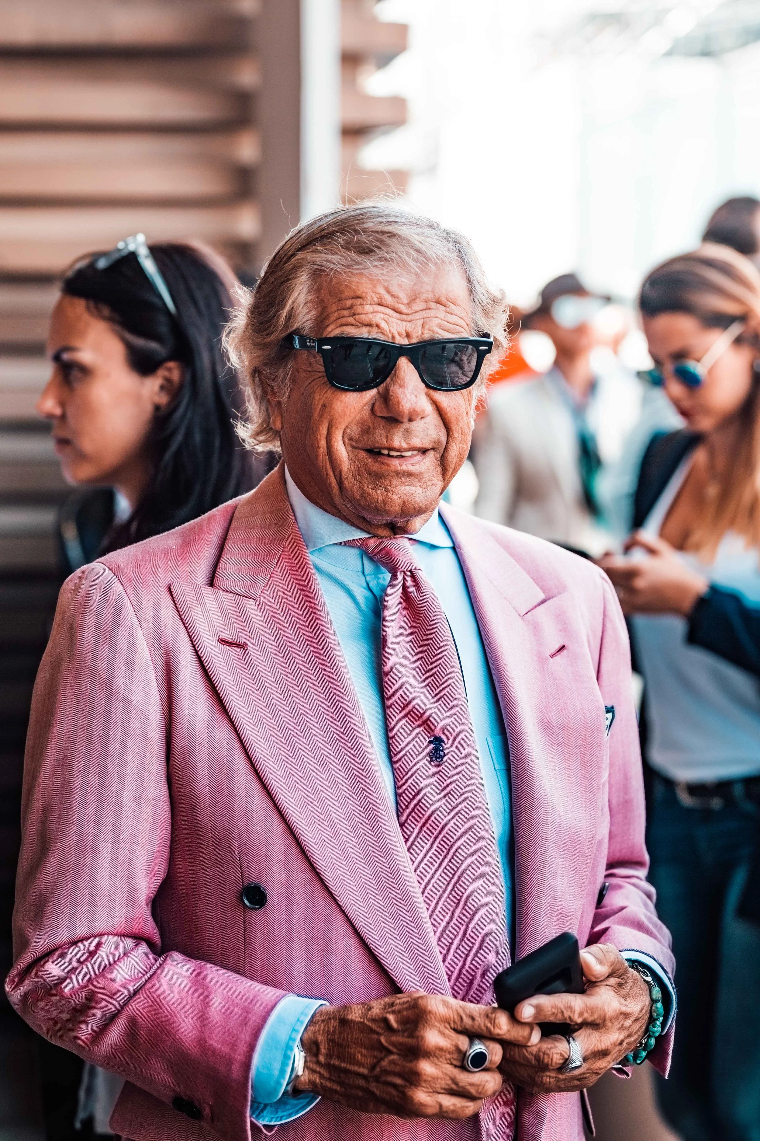 An older man wearing sunglasses and a pink suit with a matching pink tie, holding a smartphone, at an outdoor event with people in the background.