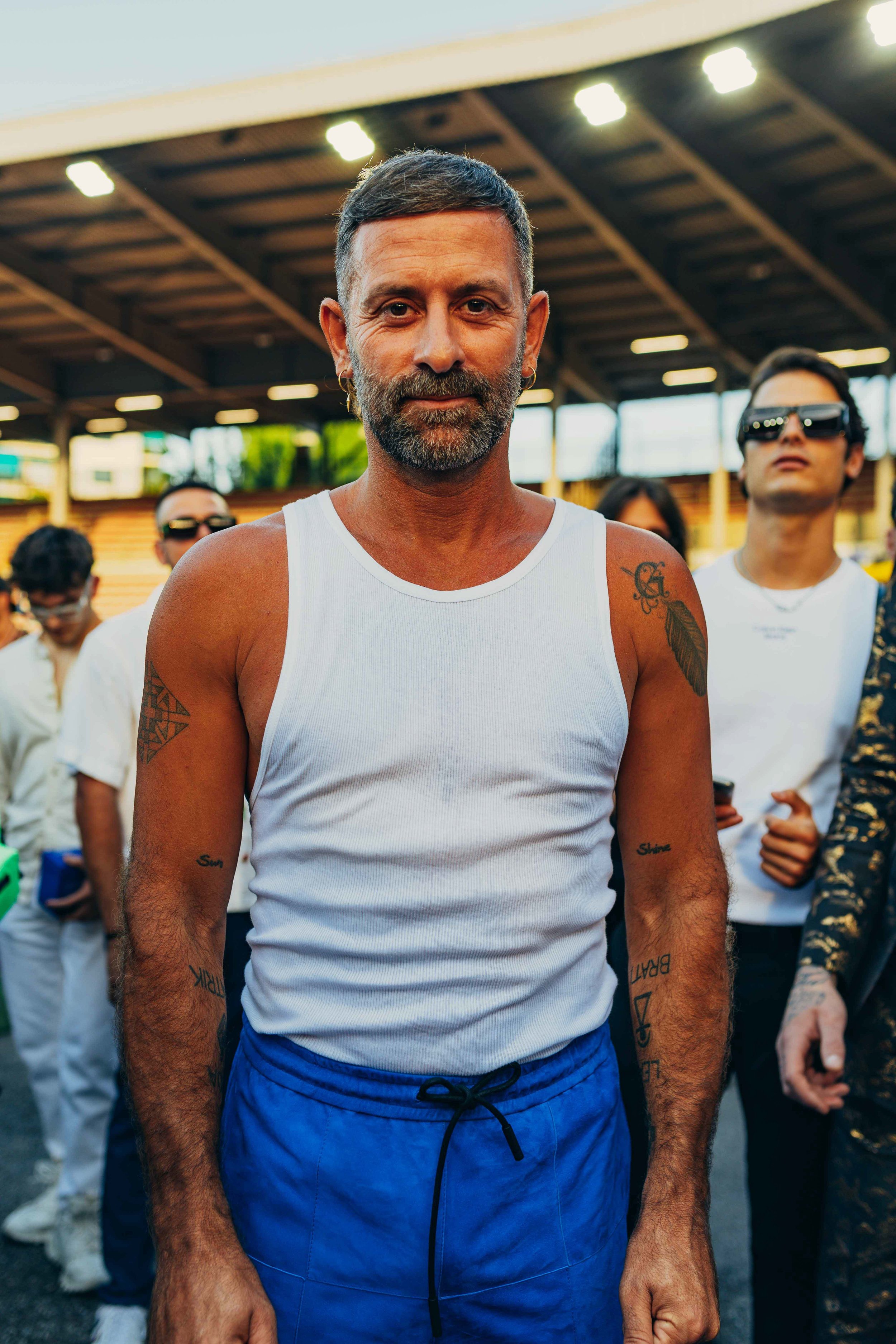 A man with a beard and tattoos, wearing a white tank top and blue shorts, stands in a group at an outdoor sports venue under a roofed structure.