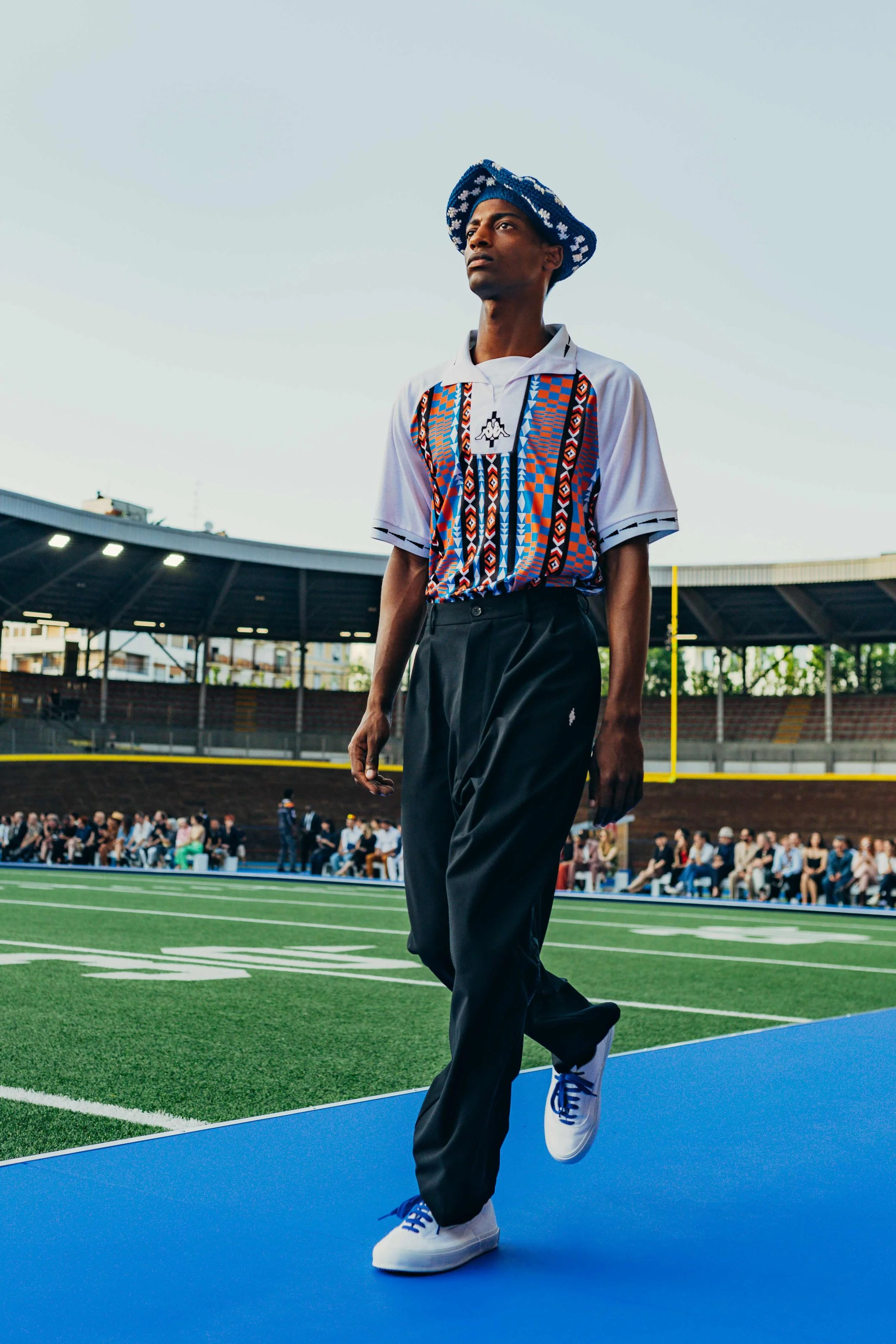 A man walking on a blue track at a sports stadium, dressed in a white and colorful patterned shirt, black pants, white sneakers, and a blue and white hat, with a crowd seated in the stands behind him.