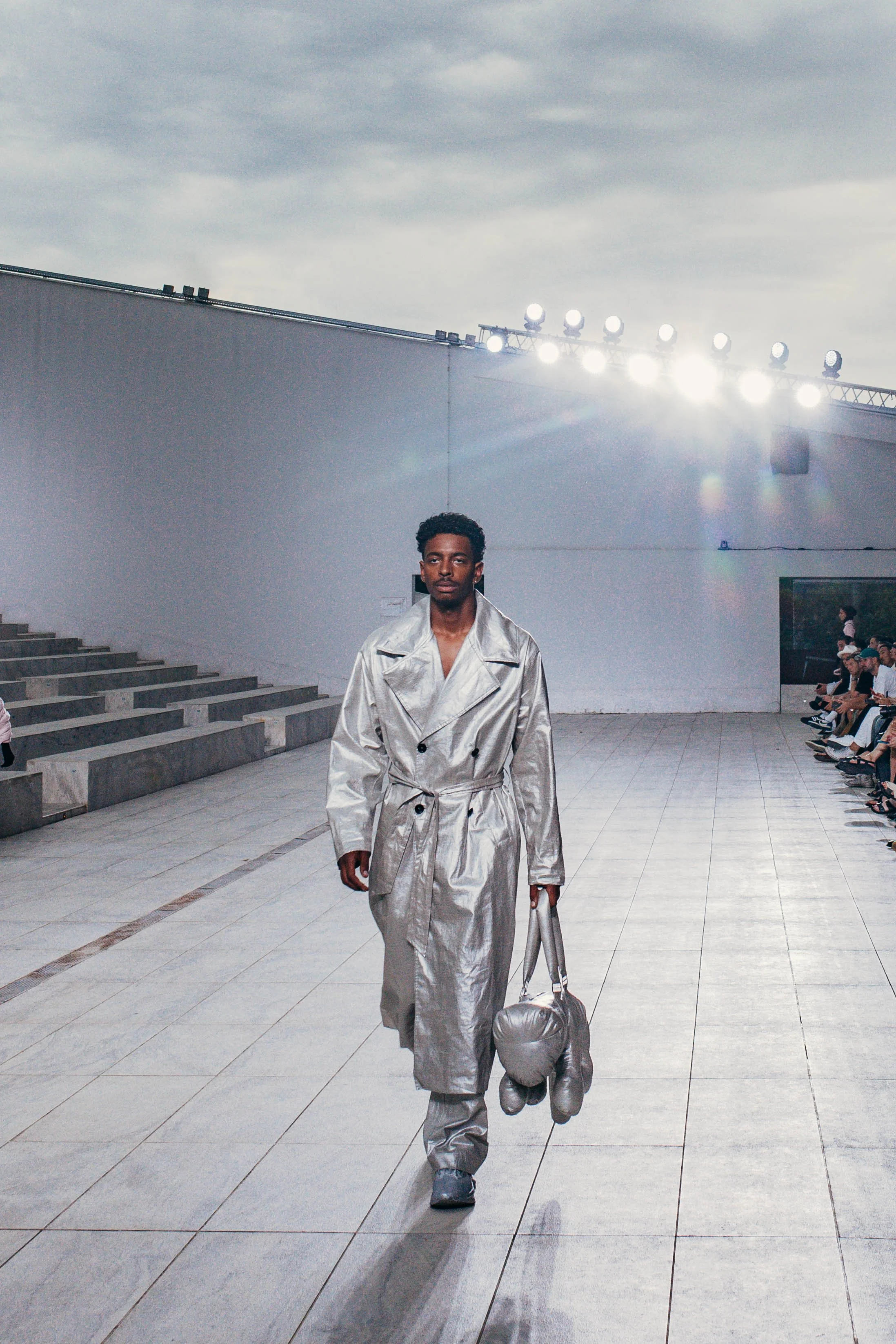 A male model walks down an outdoor runway during a fashion show, wearing a silver trench coat and pants, carrying a matching bag and boxing gloves, with runway lights above and an audience seated on the side.