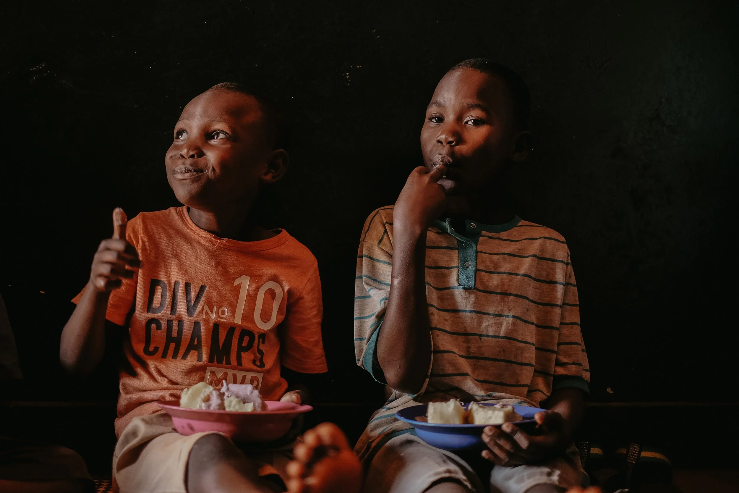 Two young boys sitting against a dark background, holding small plates with food, eating and smiling.