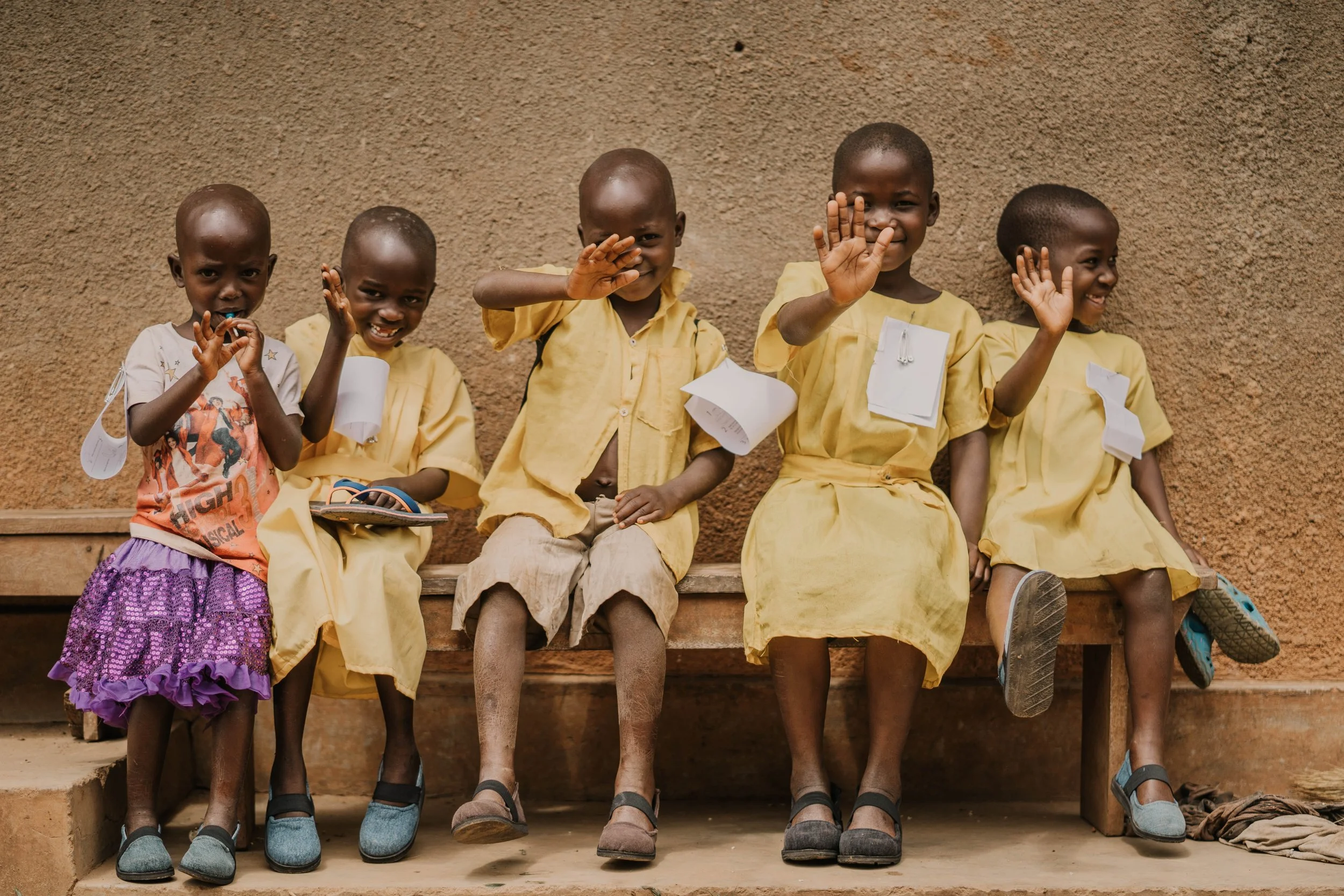 Five young children wearing Sole Hope shoes sitting on a wooden bench against a textured wall, smiling and waving at the camera. Four children are wearing yellow school uniforms, and one girl is in a purple skirt and orange shirt.