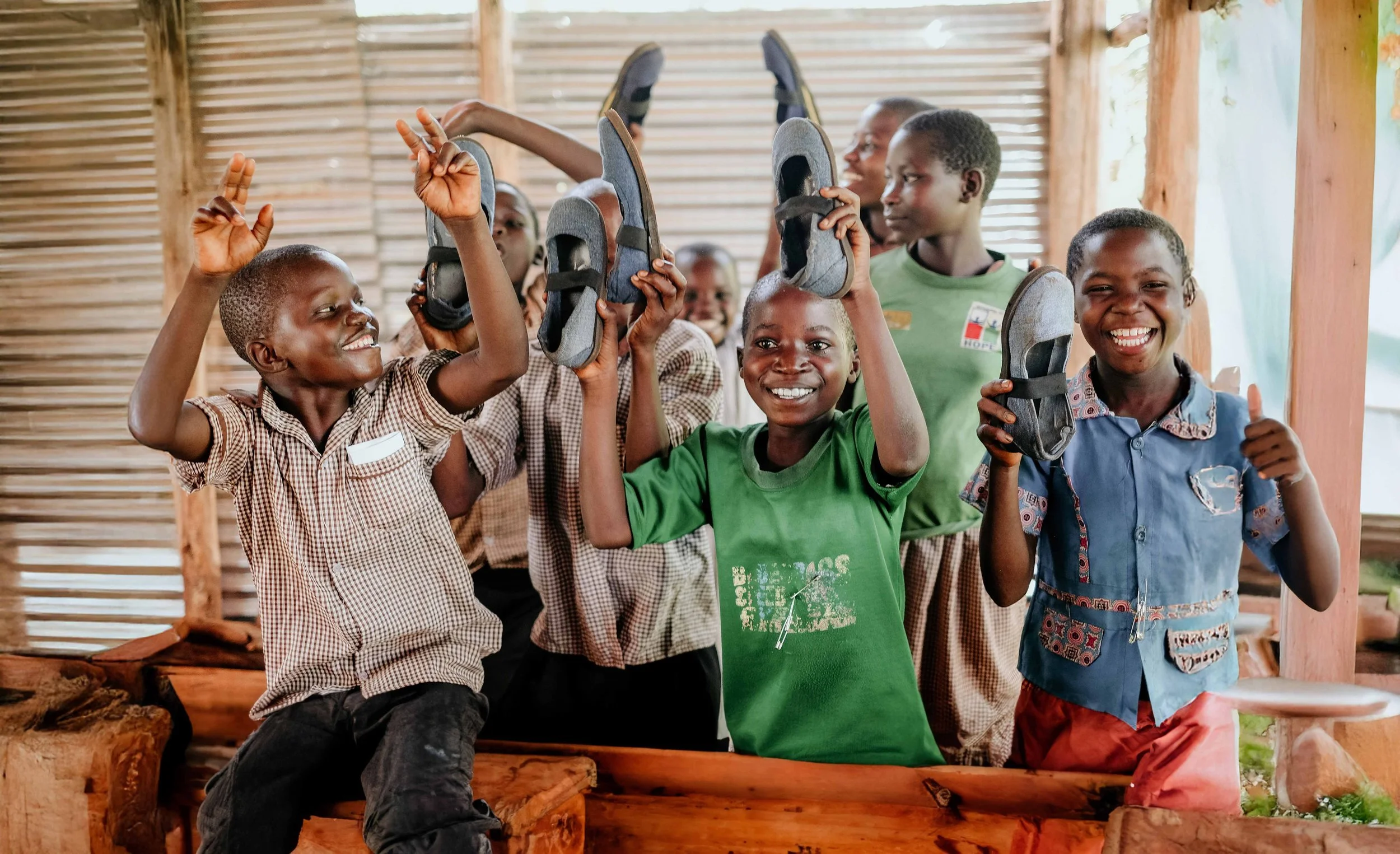 Smiling children with shoes in a wooden classroom, celebrating happily with thumbs up and raising shoes.