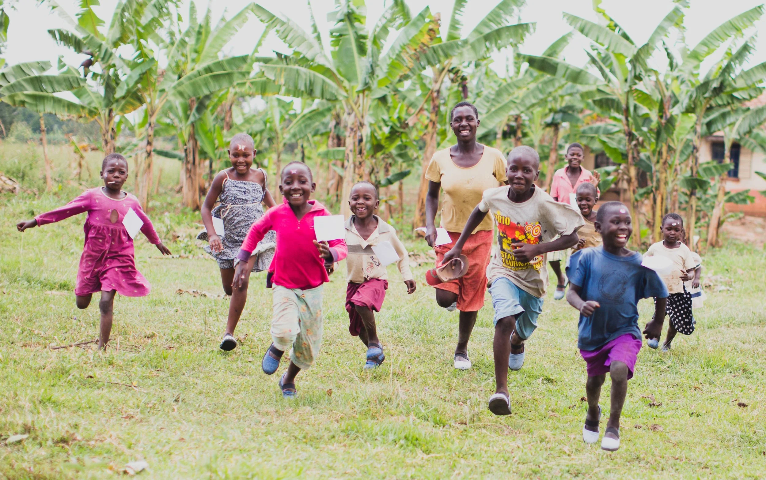 Children wearing Sole Hope shoes running happily across a grassy field in front of a banana plantation.