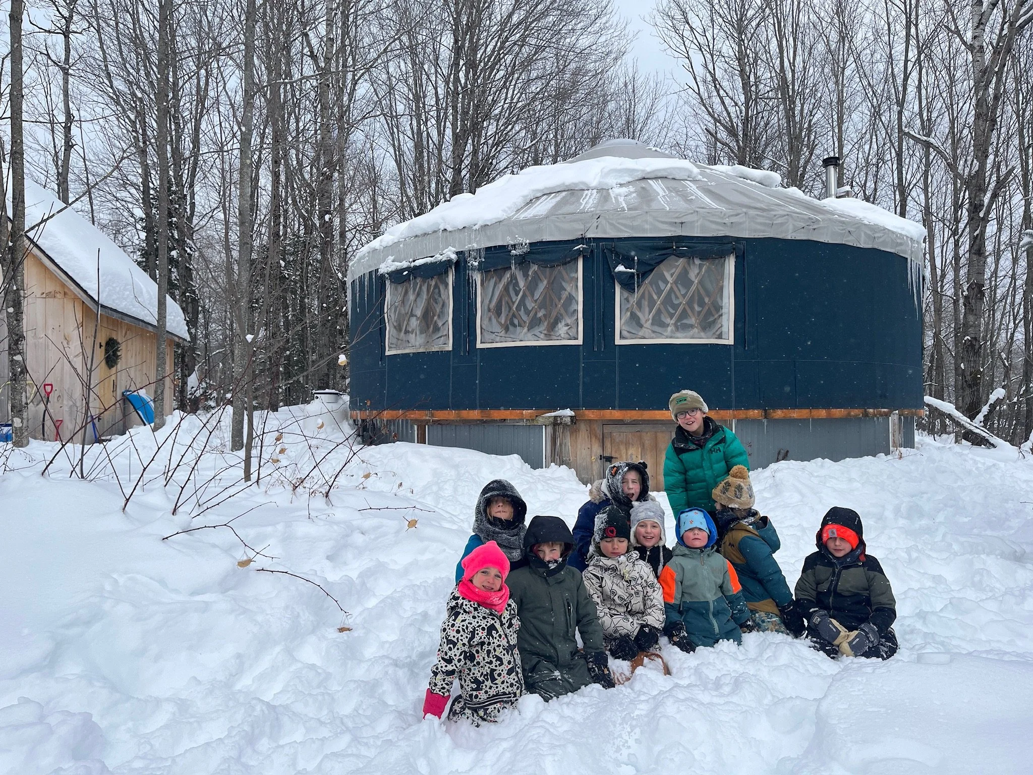 Children and adult outside in snow near a round yurt in a wooded area during winter.