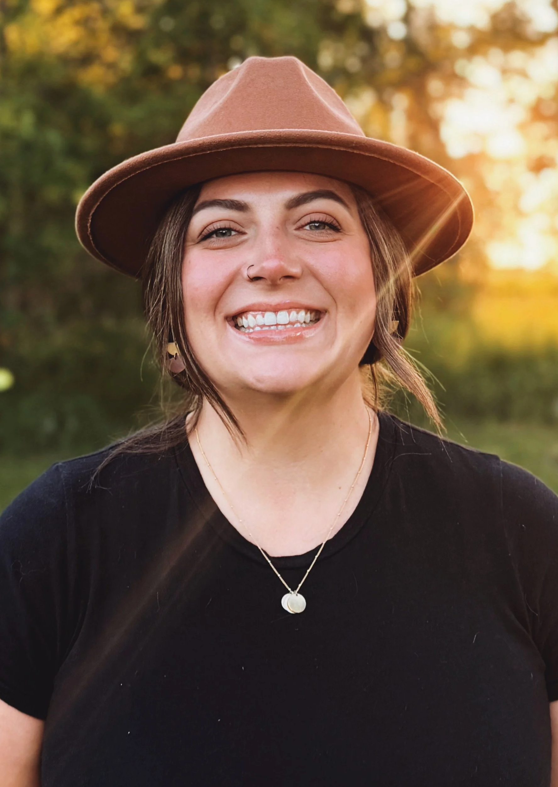 A young woman wearing a brown hat and a black top, smiling outdoors during sunset with autumn trees in the background.