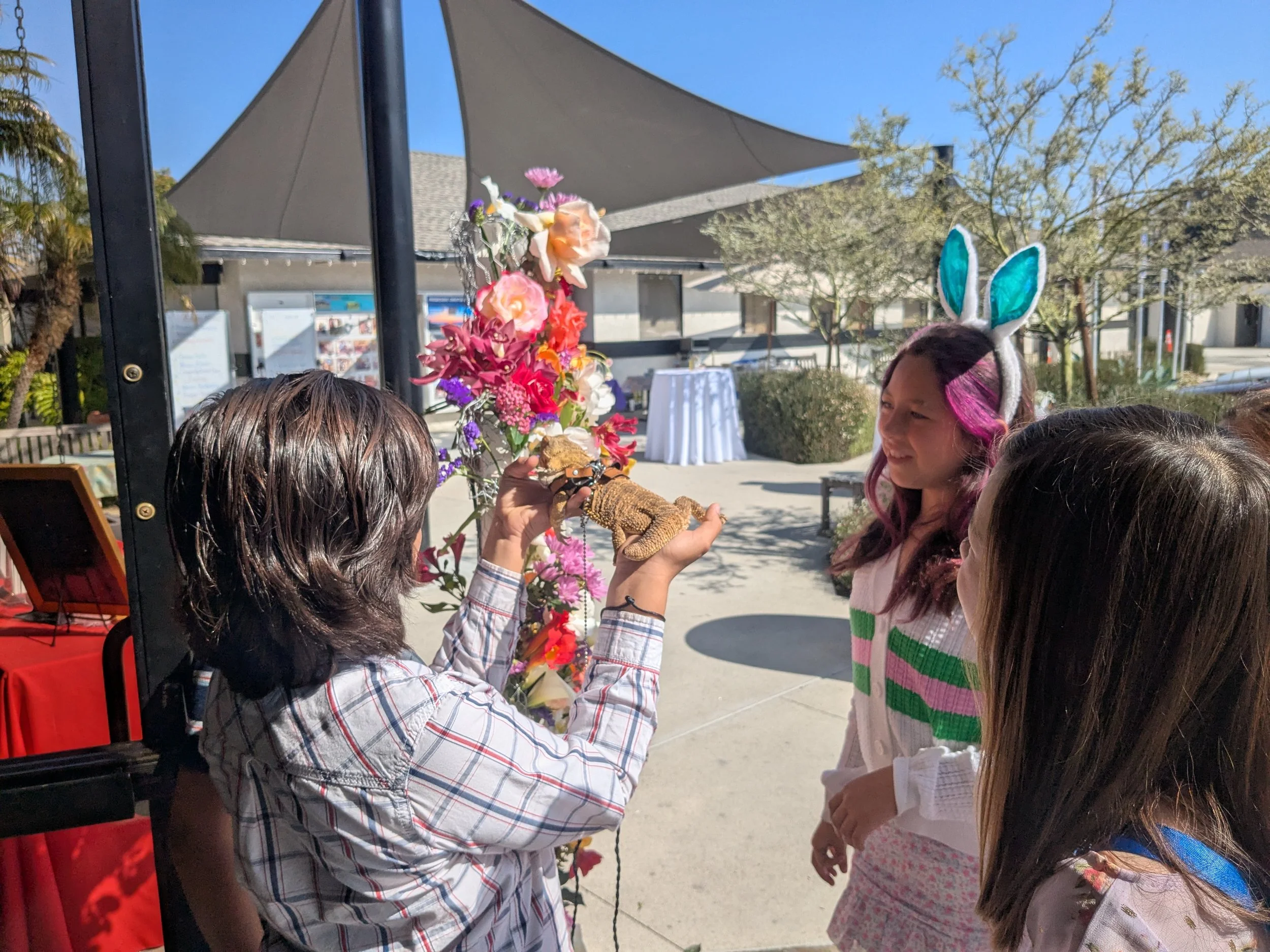 Three kids - a boy with a bearded dragon a girl in bunny ears and another girl stand by a flower cross