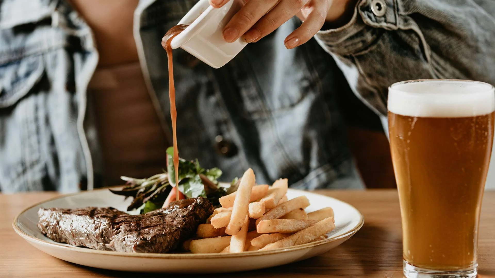 Person pouring sauce over steak with fries and salad, next to a glass of beer on a table.