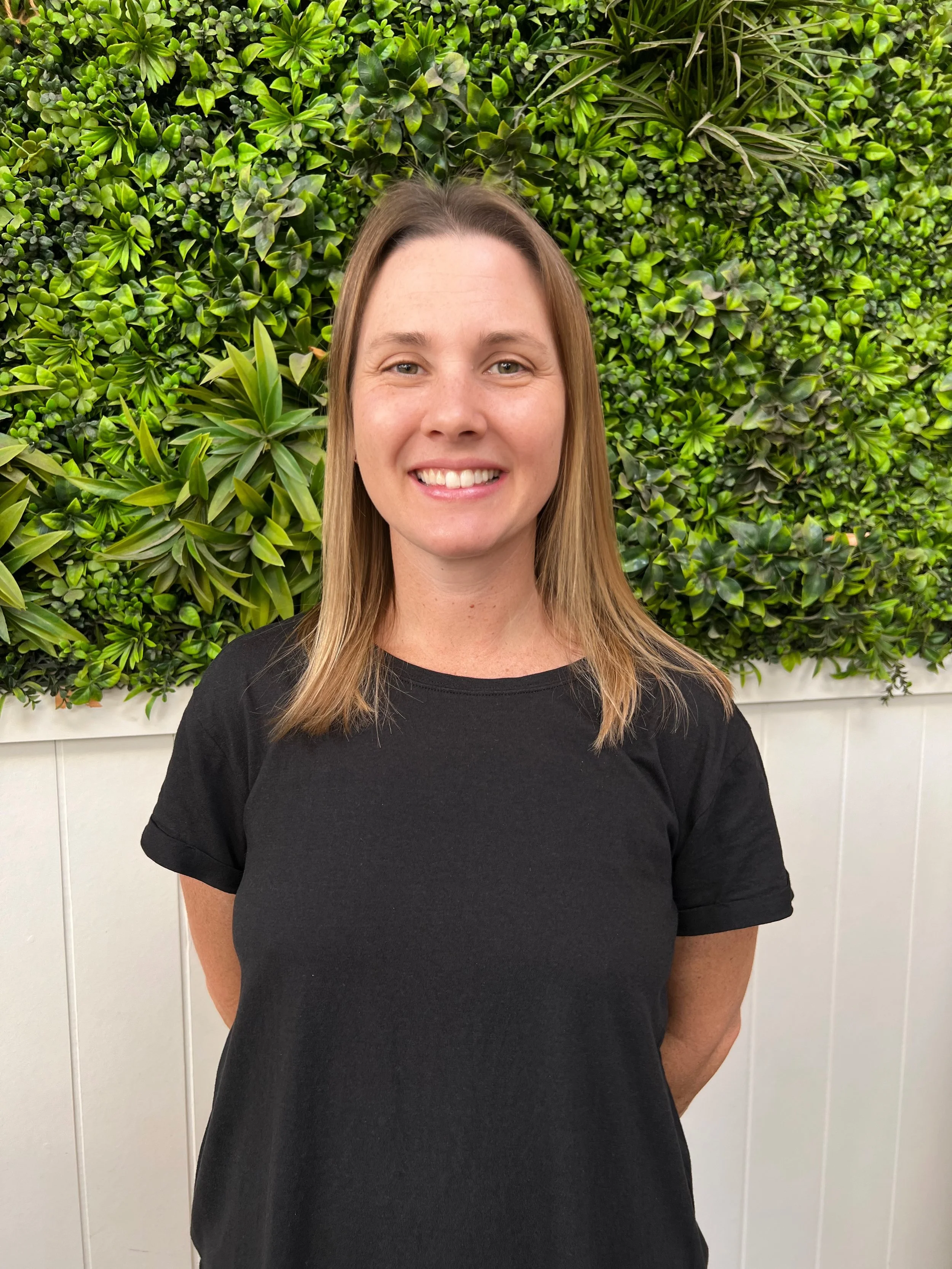 Woman standing in front of a lush green plant wall, wearing a black t-shirt, smiling at the camera.