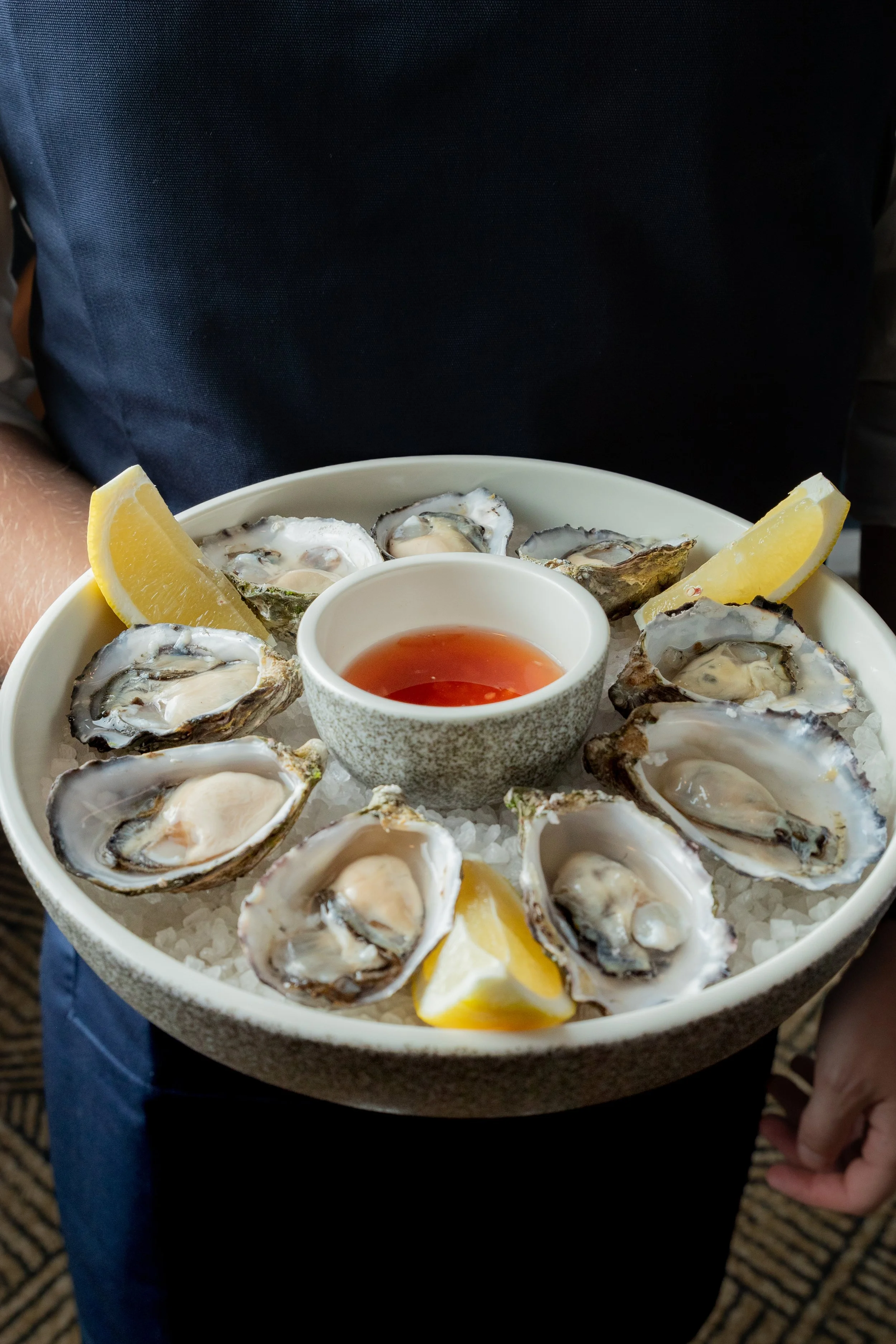 A plate of fresh oysters on ice with lemon wedges and a bowl of red sauce in the center.