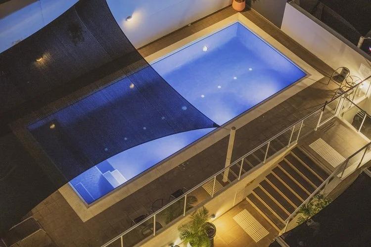 Overhead view of a modern rectangular swimming pool illuminated at night, surrounded by a tiled deck and protected by a black shade sail. The area includes lounge chairs and potted plants.