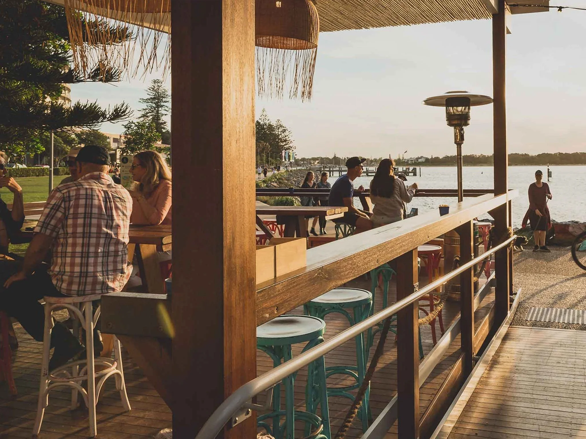 Outdoor café by the waterfront with people sitting at tables under a canopy, enjoying drinks and socializing. The area has wooden furniture and a sunny, scenic view of the water.