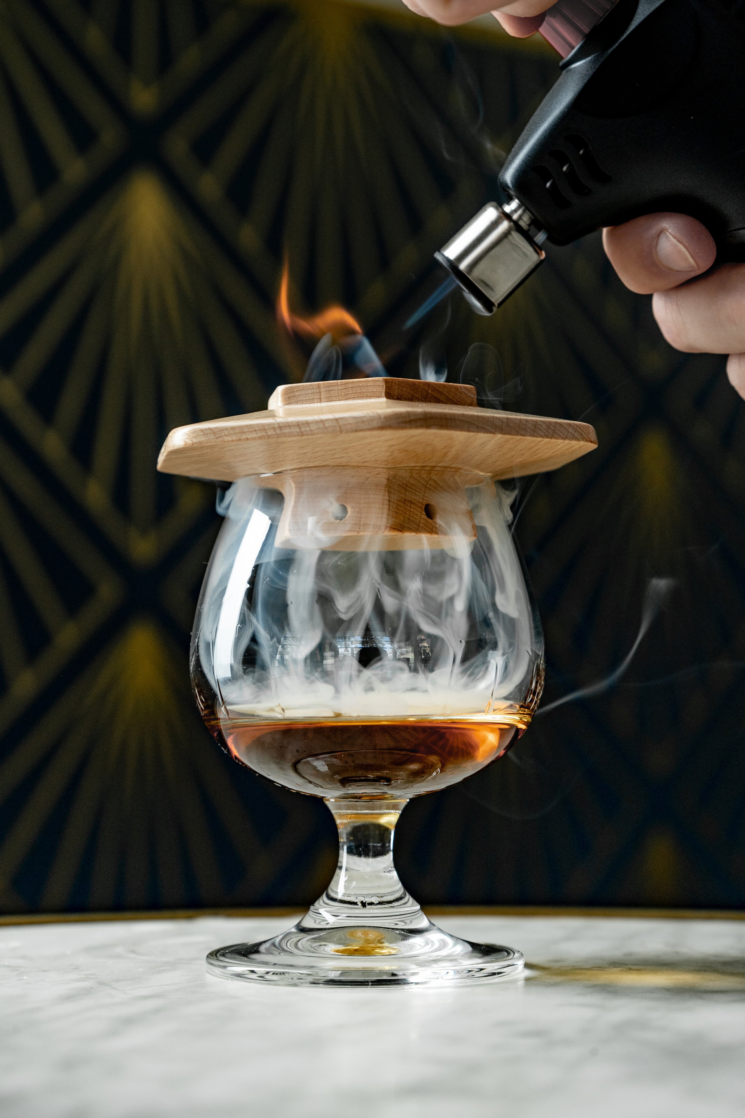 Glass of whiskey on table with smoke captured under a wooden lid while using a torch.