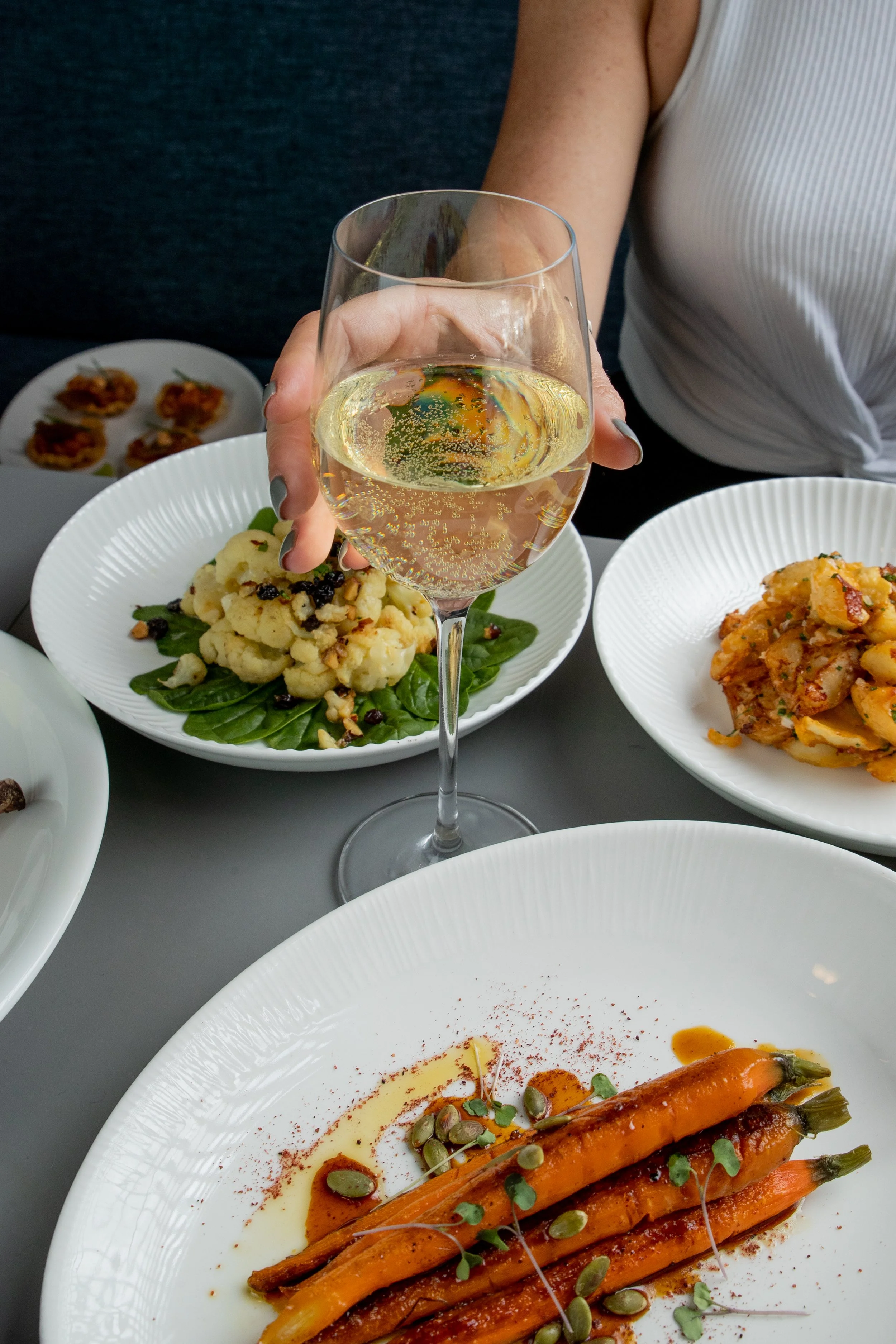 Table setting with a hand holding a glass of white wine, surrounded by dishes of roasted carrots, cauliflower salad, and pasta on white plates.