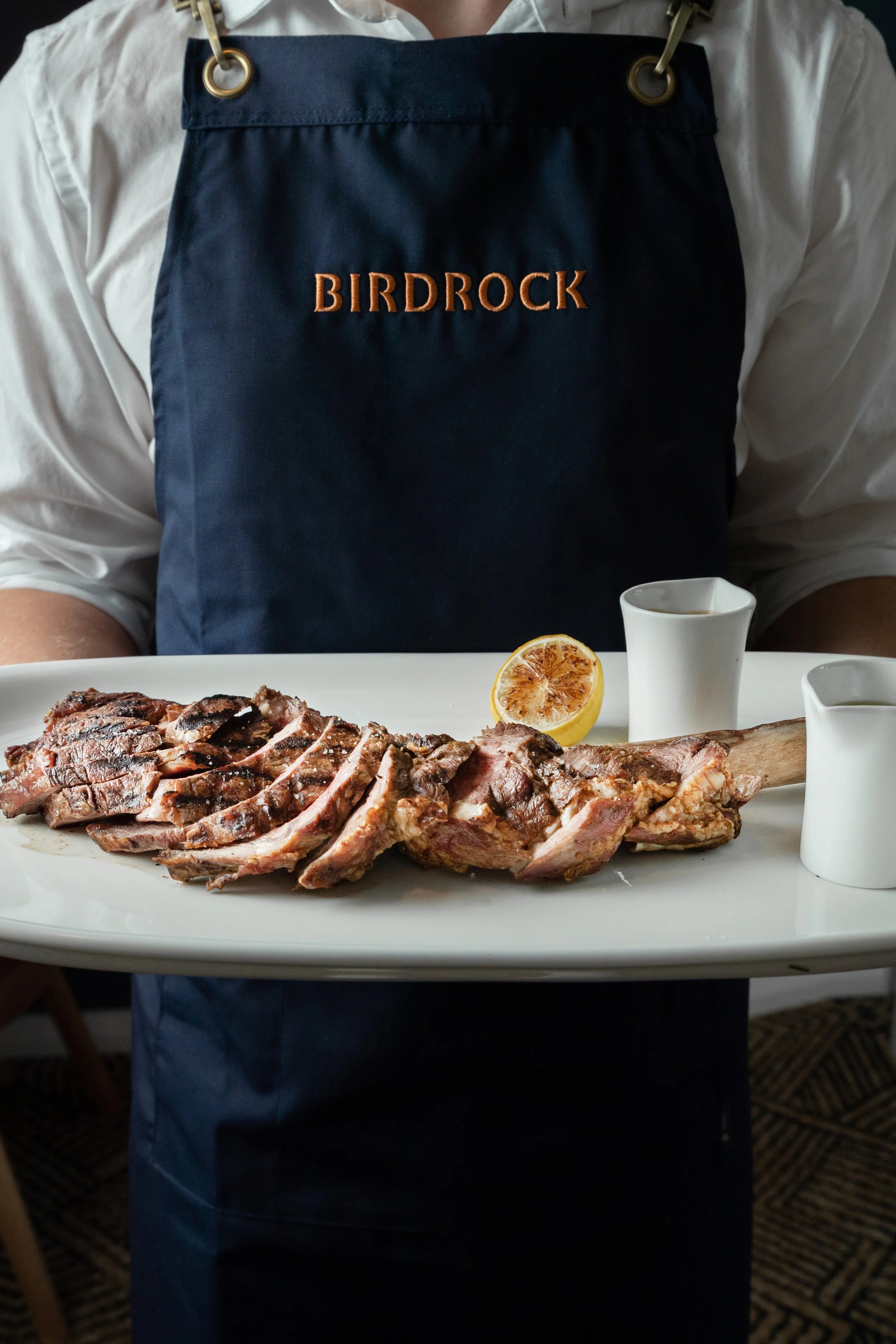 Person holding a platter with sliced grilled tomahawk steak, a grilled lemon half, and two small sauce pitchers. The person is wearing a navy apron with text "BIRDROCK."