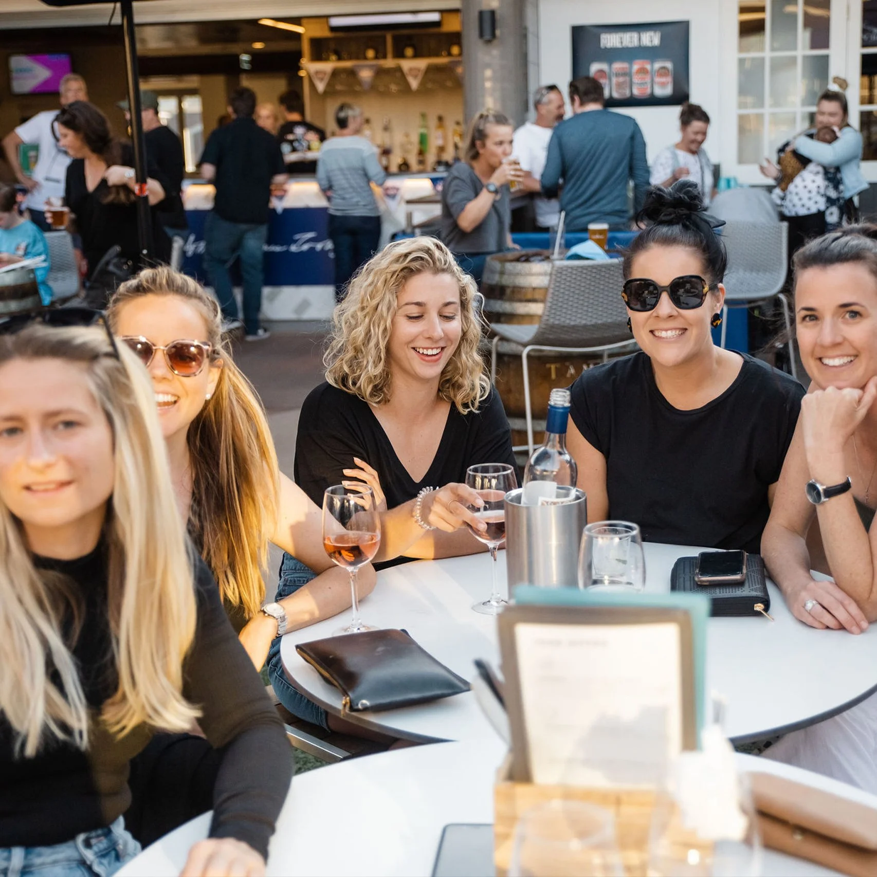 Group of women sitting at a table outdoors, smiling and enjoying drinks. Other people are in the background at a bar area.