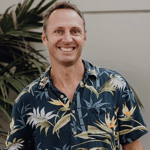Smiling person wearing a tropical floral shirt with palm leaves in the background.