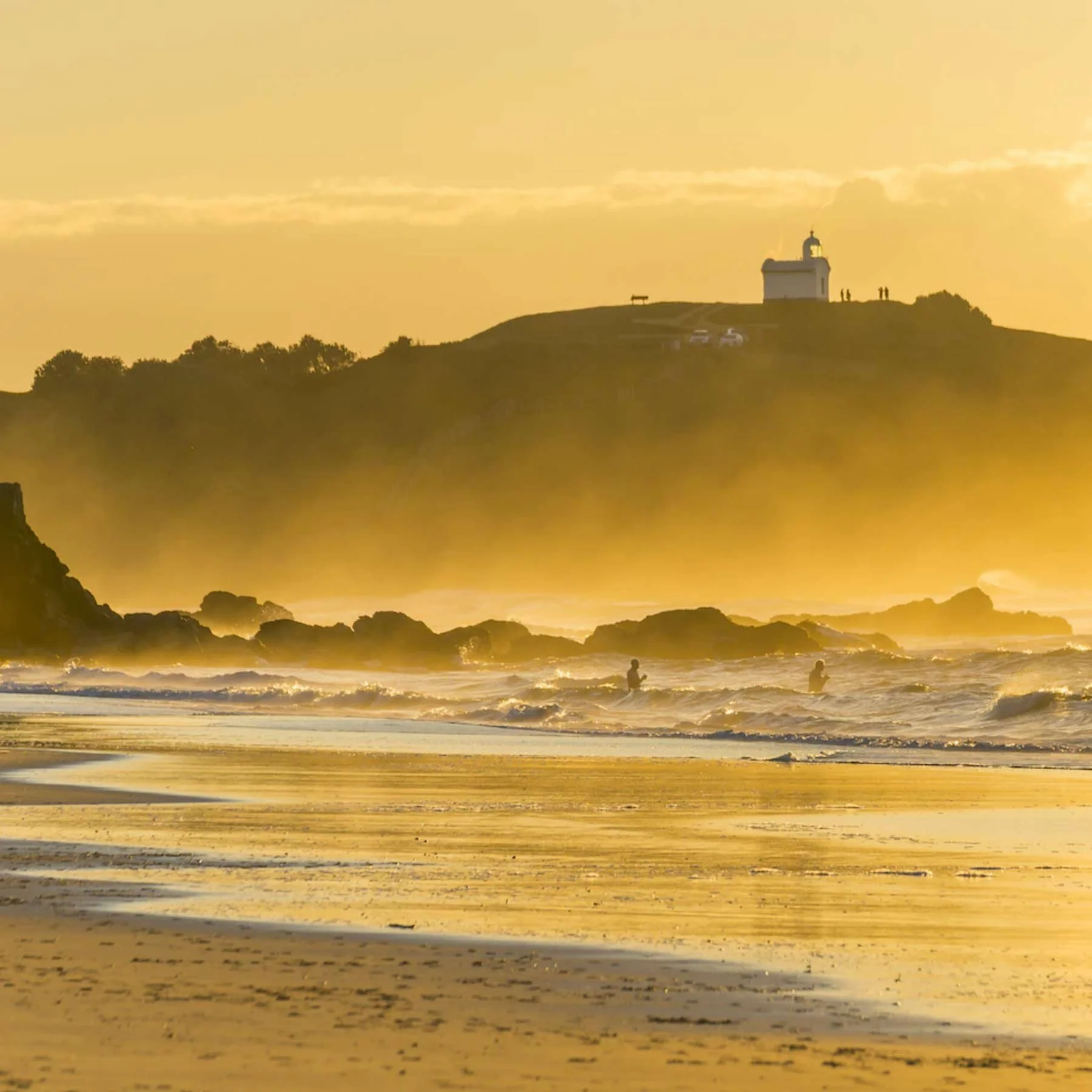 Sunset beach scene with surfers in the water, rocky shoreline, and a small white building on a hill in the background.