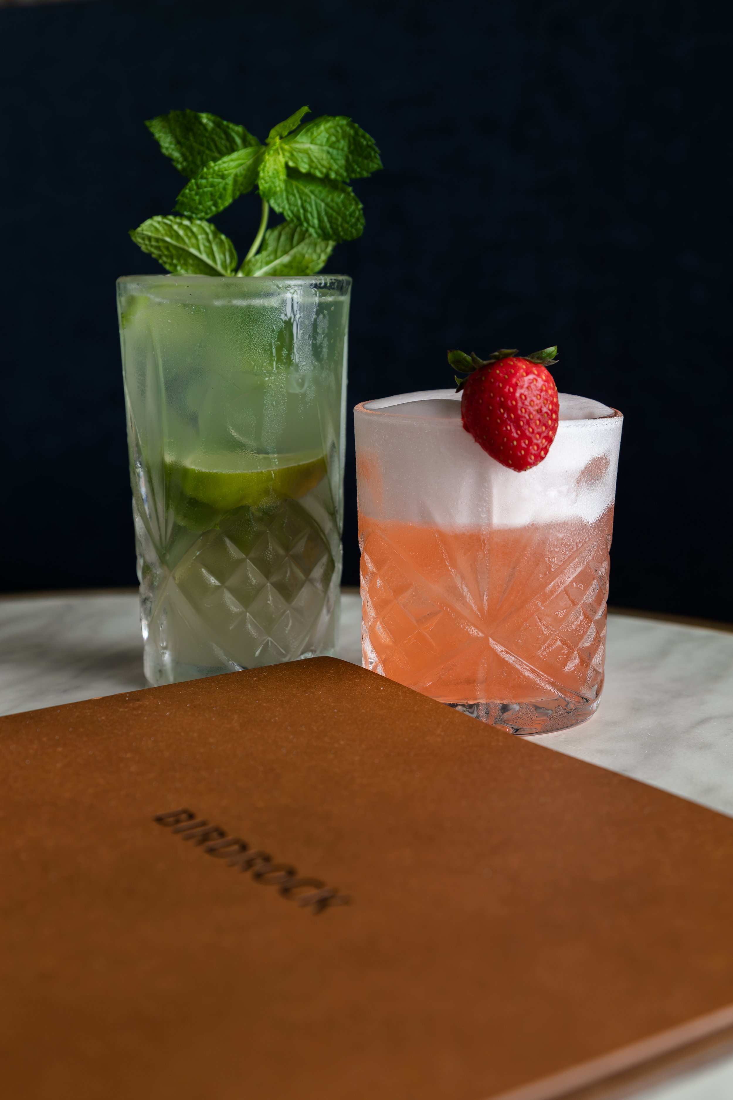 Two cocktails on a table; one is green with mint and lime garnish, the other is pink with a foamy top and a strawberry garnish. A brown menu with the word "BIRDROCK" is in the foreground.