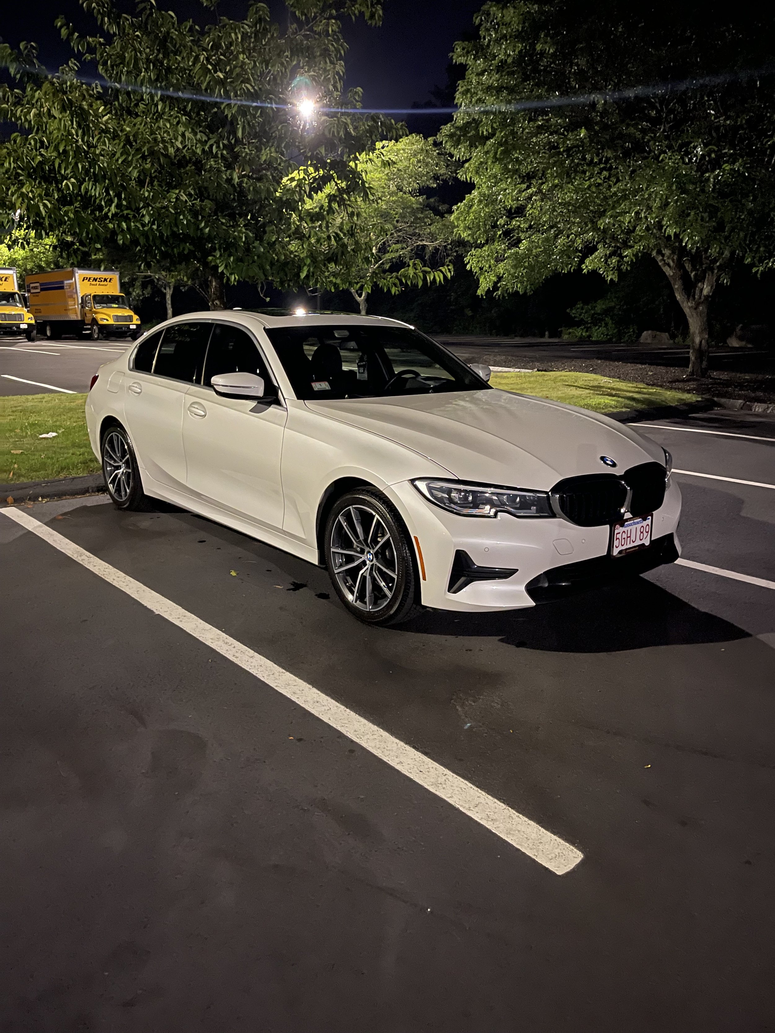 White BMW sedan parked in a parking lot at night, with trees and yellow Penske trucks in the background, illuminated by streetlights.