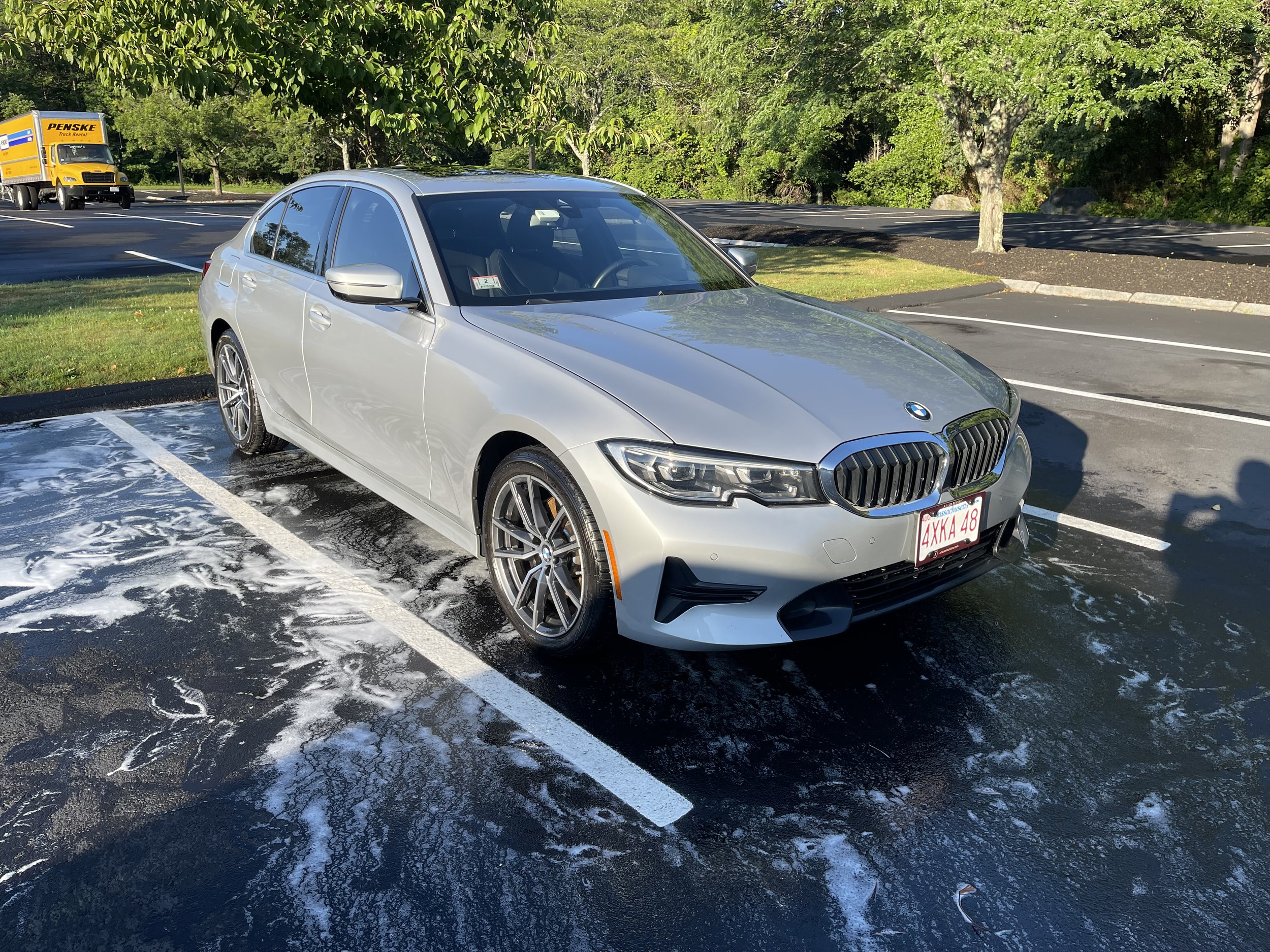 Silver BMW car in parking lot with soapy water on the ground; yellow Penske truck in the background.