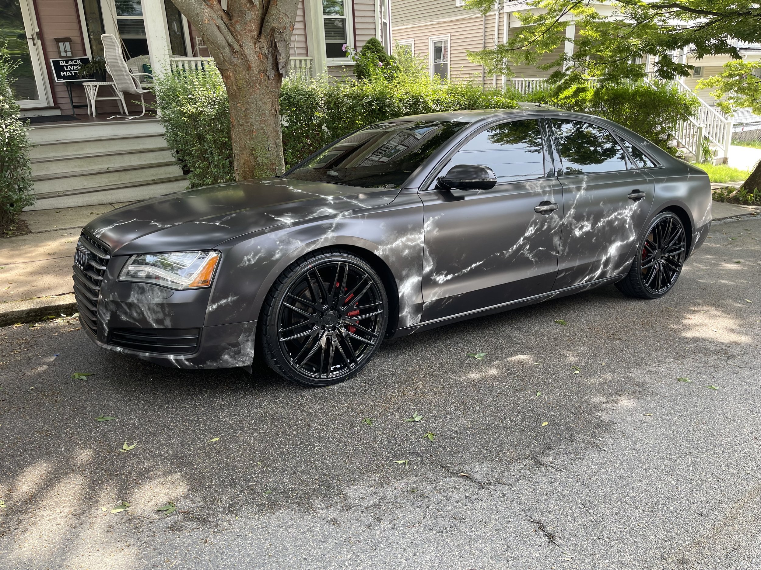 A customized car with a matte gray and marbled paint job, black rims, and parked on a residential street. A "Black Lives Matter" sign is visible on a porch in the background.
