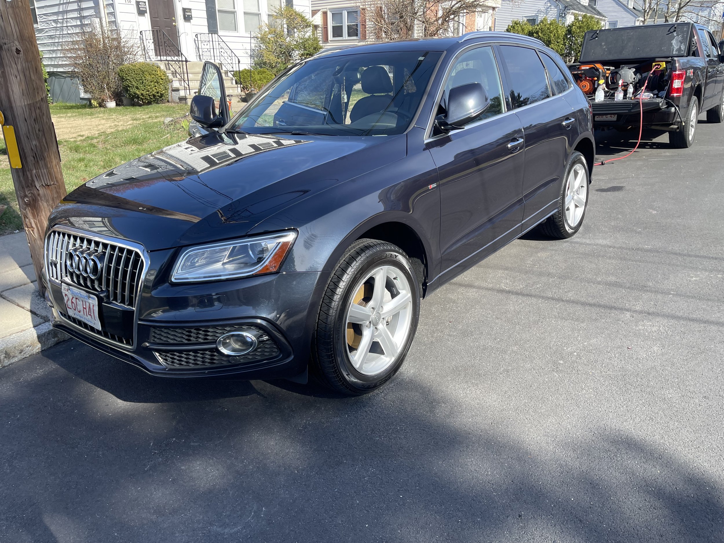 A dark-colored Audi SUV parked on the street with a pickup truck in the background. The Audi is facing slightly to the right, with houses visible in the distance.