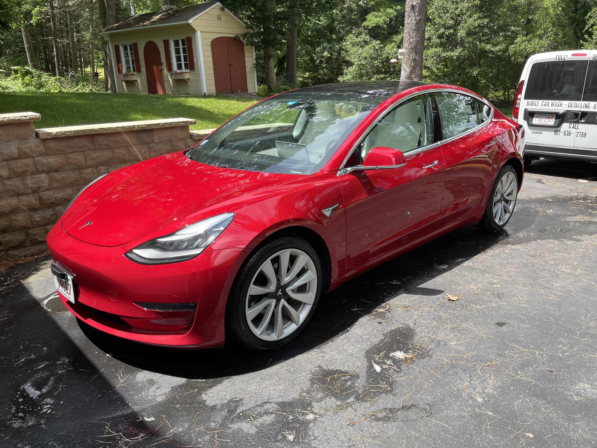 Red Tesla Model 3 parked on a driveway next to a van and a garden shed.