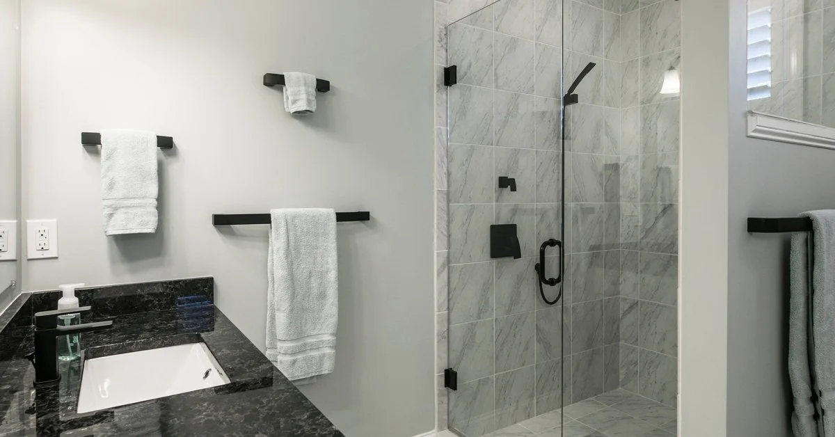 A shower with gray tiles has a glass a pivot glass door. The bathroom vanity is black, and the space has black fixtures.