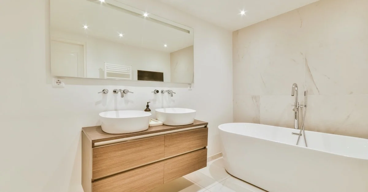 A small, double sink vanity with a floating, frameless mirror. The rest of the bathroom has white tiling and a free-standing tub.
