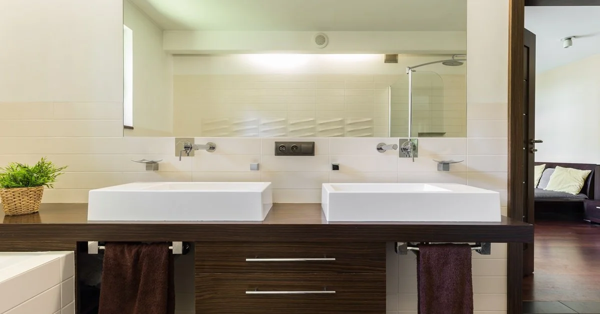 A master bathroom with brown tiles and a brown wooden vanity with double sinks. There is a long, frameless mirror above it.