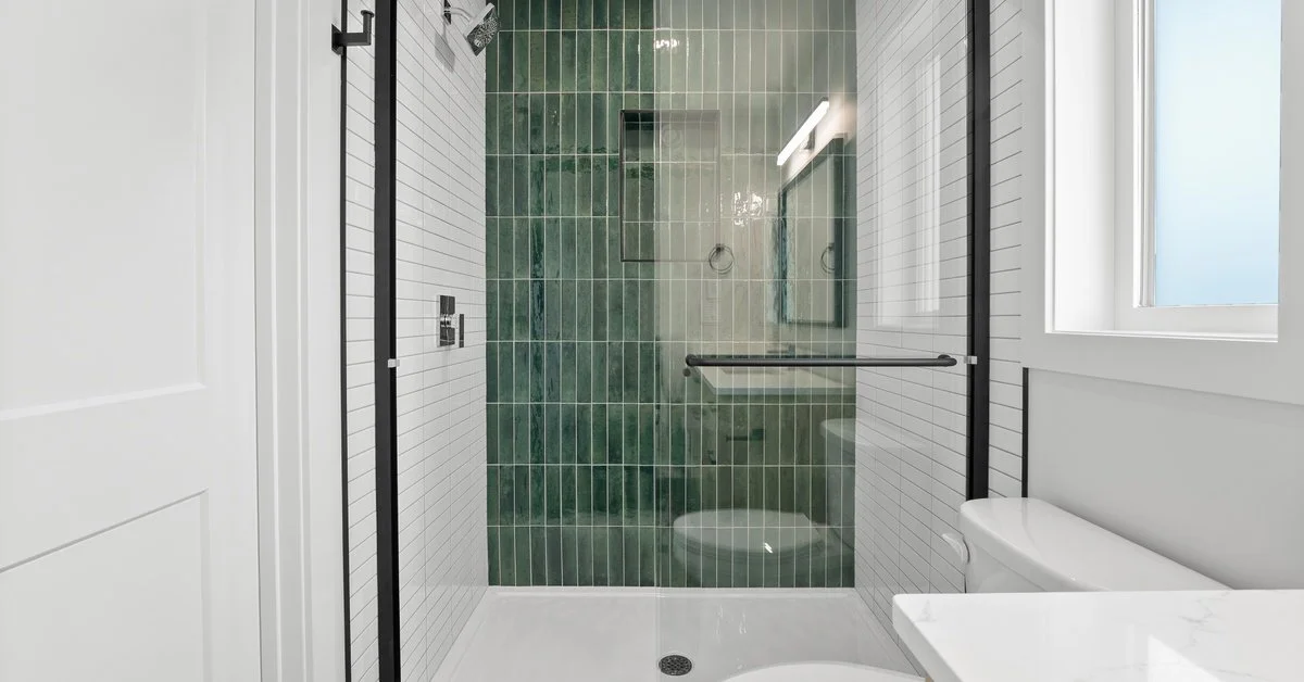 A shower with white tile floors and walls has one green accent wall and a black frame around the glass shower door.